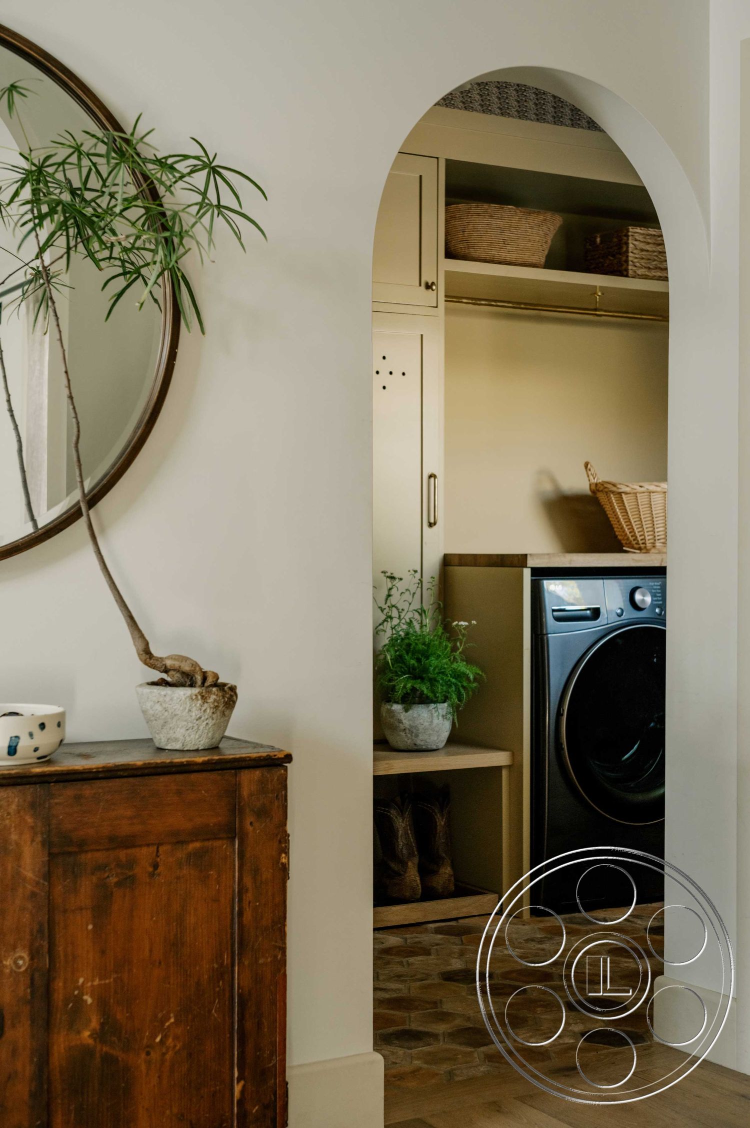 Americana 429 - natural wood accents, minimalist design elements, soft color palette, contemporary interior style, open layout design, arched entryway interior, hexagon tile flooring in laundry area, wooden storage cabinet with vintage finish, round mirror wall decor, textured wall paint in neutral color, overhead shelf with woven baskets, indoor potted plant decor, laundry room with washer and dryer, multifunctional storage solutions, warm lighting fixtures
