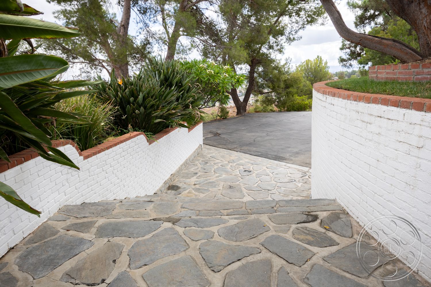 Sinatra House - exterior, paved driveway, lush greenery, suburban setting, natural stone steps, flagstone pathway, textured stone surface, landscaped garden area, white painted brick wall, red brick border, curved brick wall, earth-toned landscaping
