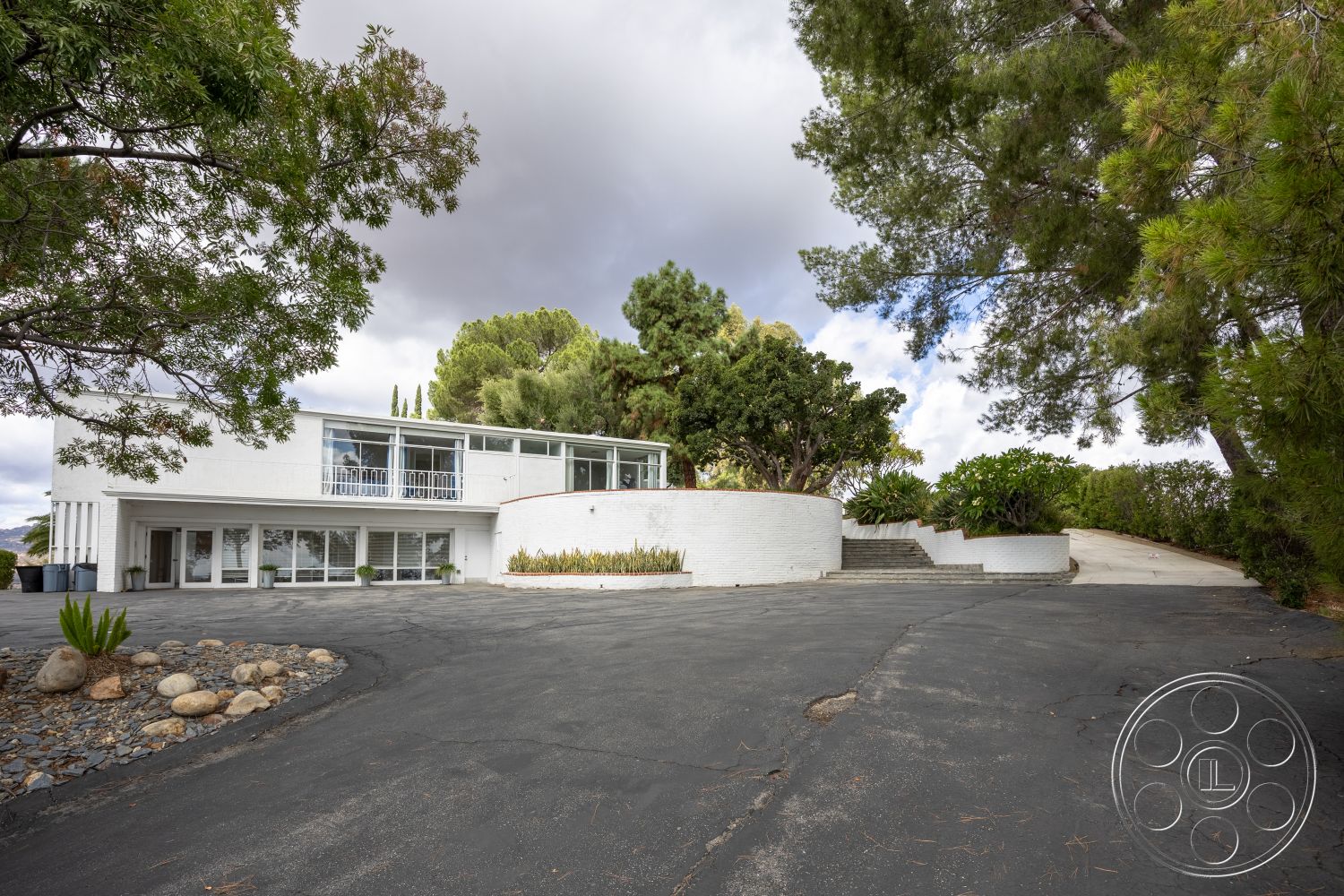 Sinatra House - parking, outdoor seating area, curved walkway, asphalt driveway, floor-to-ceiling windows, city skyline view, hillside location, sloped terrain, surrounded by lush greenery, rectangular patio area, mid-century modern architectural style, exterior white stucco walls, exposed concrete steps, metal handrails, curved entryway, concrete planter boxes, stone garden border, landscaped patio area, natural stone accent landscaping, paved side driveway