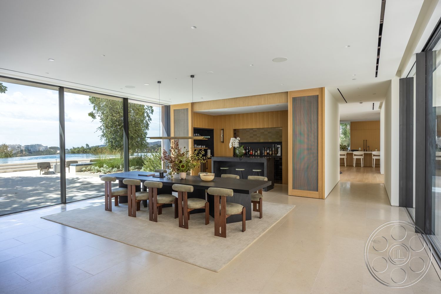 Modern 511 - interior dining room with large glass windows, stone floor tiles in light cream color, wooden dining table with upholstered chairs, contemporary eat-in kitchen with sleek cabinetry, open floor plan layout with seamless indoor-outdoor flow, neutral color palette with soft lighting fixtures, modern bar area with dark stone countertop, wood accent wall in kitchen with minimalist decor, large format glass sliding doors leading to patio, textured beige area rug under dining table, architectural feature of vaulted ceiling with recessed lighting, surrounding views of waterfront landscape, natural light flooding from floor-to-ceiling windows, minimalist modern design with functional aesthetics