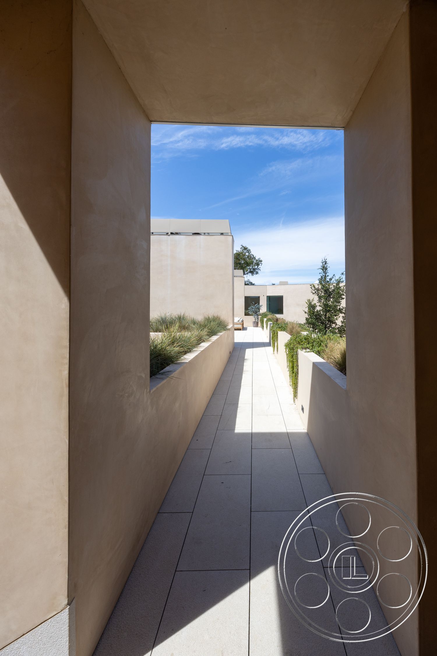 Modern 511 - exterior, modern aesthetic, textured plaster walls, stucco wall finish, lush garden setting, minimalist architectural style, natural light emphasis, clean lines and geometric shapes, rectangular walkway, large format stone pavers, landscaped edges with greenery, sunlit corridor, open-air passage, vibrant blue sky view, soft sunlight shadows, narrow pathway design