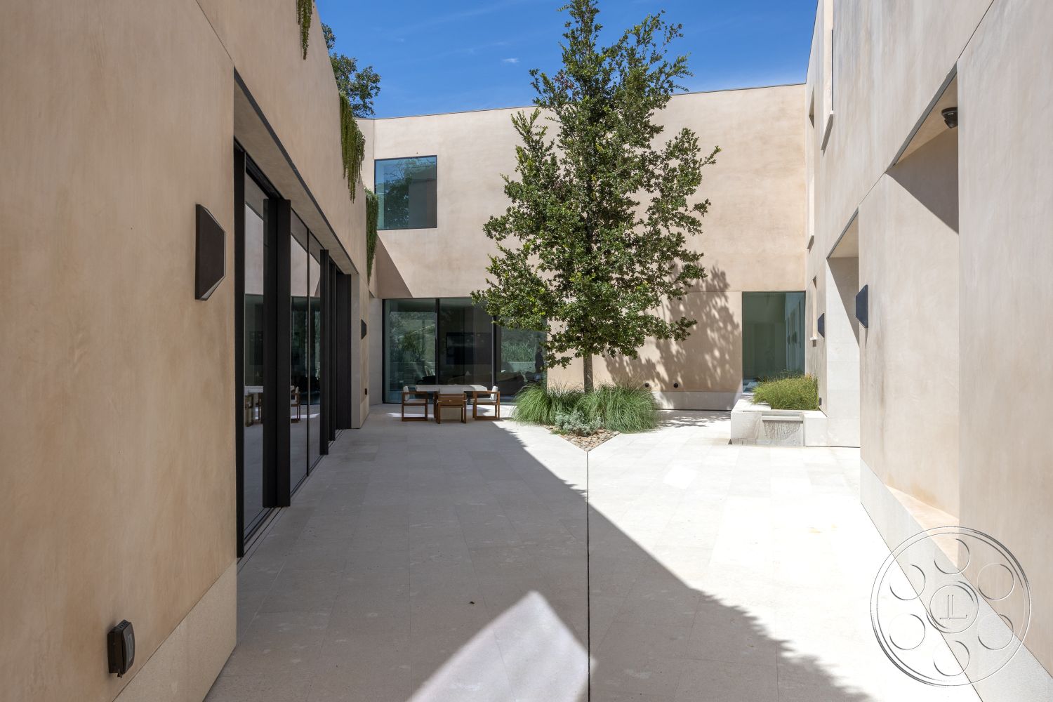 Modern 511 - large glass sliding doors, contemporary architectural style, exterior courtyard with smooth stucco walls, concrete paver flooring in light beige, minimalist wooden outdoor furniture, mature trees integrated into landscaping, open-air design with natural light, windowed wall sections providing views, subtle earthy color palette, low-maintenance landscaping with grass and stones, urban setting with modern design elements, square courtyard layout surrounded by walls, soft afternoon sunlight creating shadows, natural materials with a focus on simplicity, inviting outdoor space for relaxation