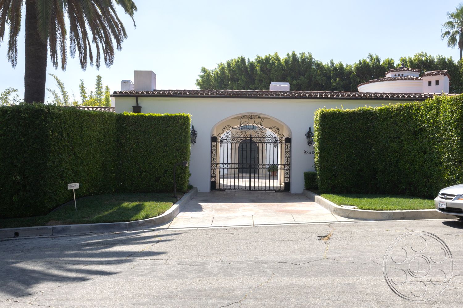 Spanish 168 - exterior, paved driveway, outdoor lighting, curved driveway, Spanish Revival architecture, stucco walls, arched entryway, terracotta tile roof, ornate wall sconces, landscaped hedges, wrought iron gate, concrete patio area, palm trees in garden, lush green lawns