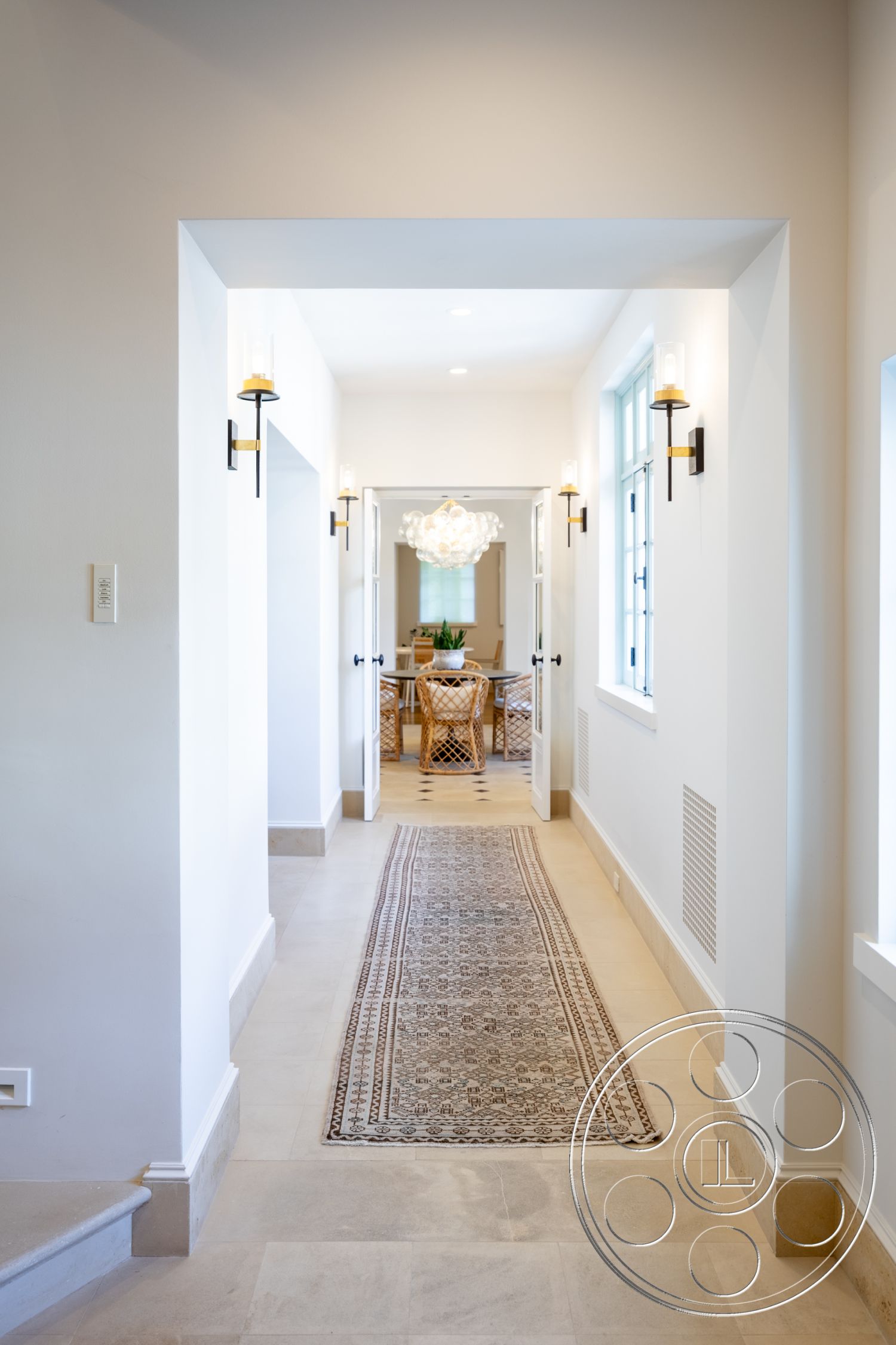 Spanish 168 - textured wall finish, soft lighting fixtures, minimalist architectural style, interior hallway with neutral painted walls, natural stone flooring in hallway, decorative hallway runner rug, exposed light fixtures with black metal accents, arched doorways leading to dining area, floor-to-ceiling windows allowing natural light, open-concept dining area visible from hallway, subtle crown molding details, defined entry with clear sight lines