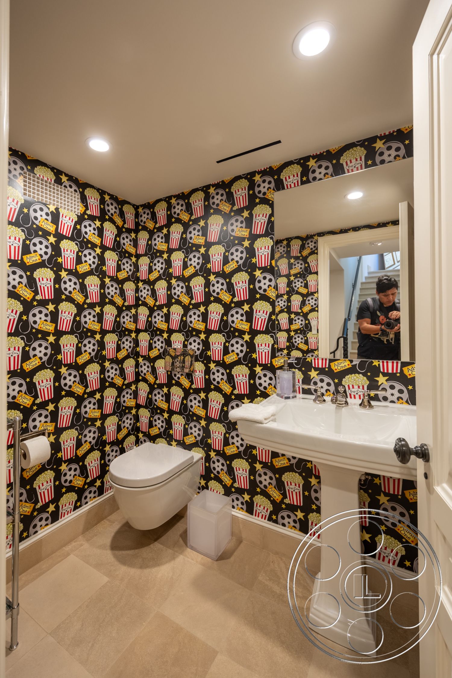 Spanish 168 - ceramic tile flooring, textured plaster walls, interior bathroom, recessed LED ceiling lights, square mirror above sink, minimalist bathroom accessories, graphic wallpaper featuring popcorn and film reels, modern pedestal sink, chrome toilet paper holder, white paneled door, neutral beige wall tiles