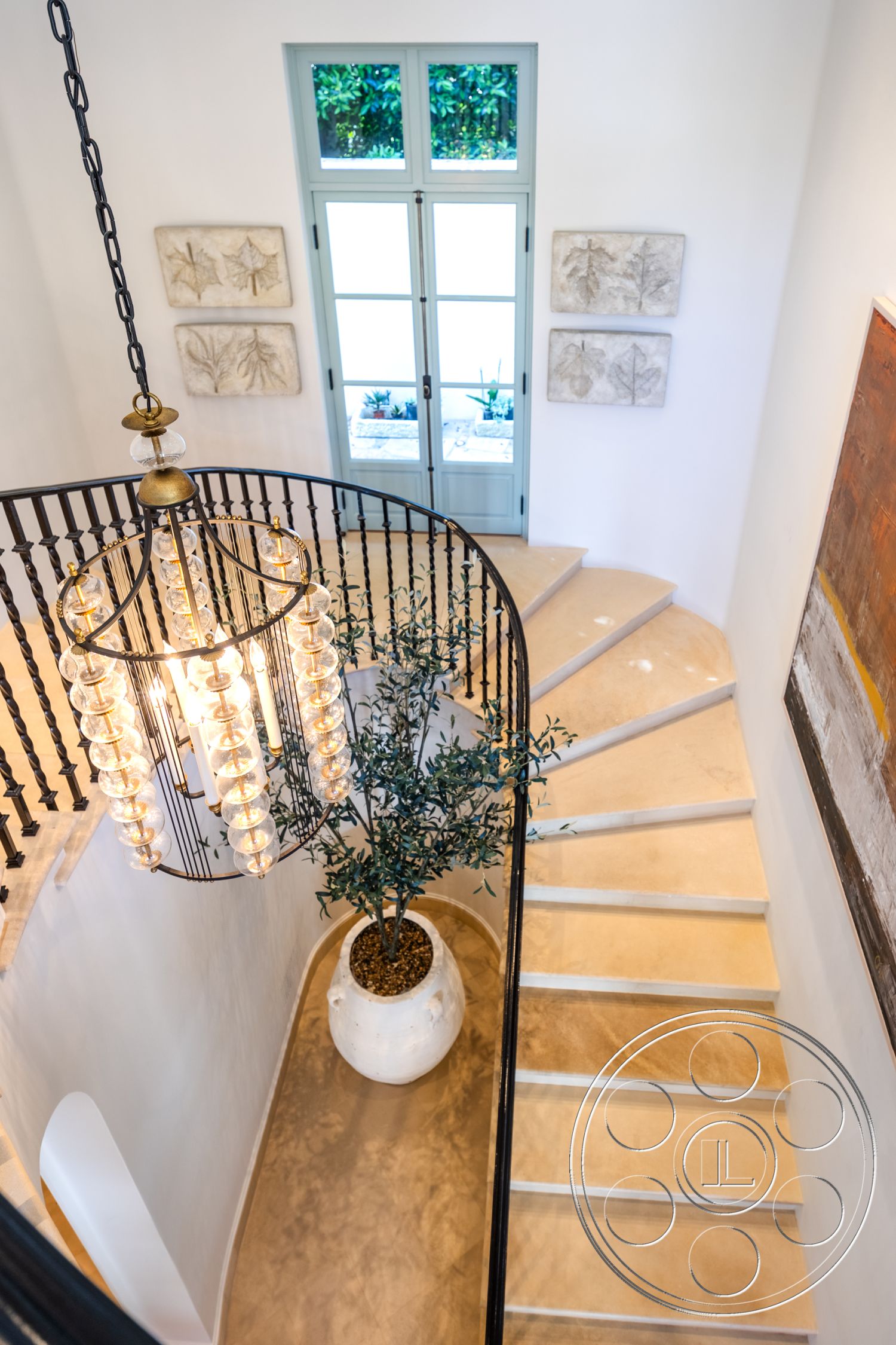 Spanish 168 - minimalist decor, natural light from large windows, arched doorways, curved staircase, floor-to-ceiling glass doors, exposed concrete staircase steps, black wrought iron staircase railing, chandelier with glass bulbs, smooth plastered interior walls, light-colored polished concrete flooring, decorative wall art panels, green indoor potted plant, white ceramic planter, modern interior design style, contemporary entryway