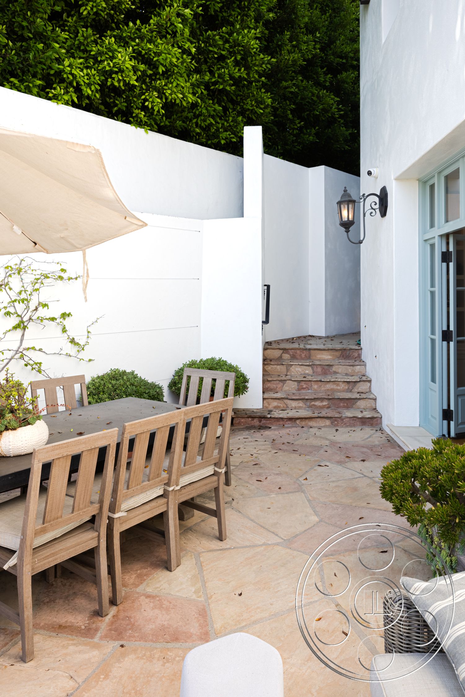 Spanish 168 - white stucco walls, minimalist design, wooden dining table, landscaped greenery, outdoor dining area, sunshade umbrella, stone steps, rustic outdoor setting, potted shrubs, exterior patio space, flagstone patio flooring, lantern-style wall sconce, surrounding wooded area