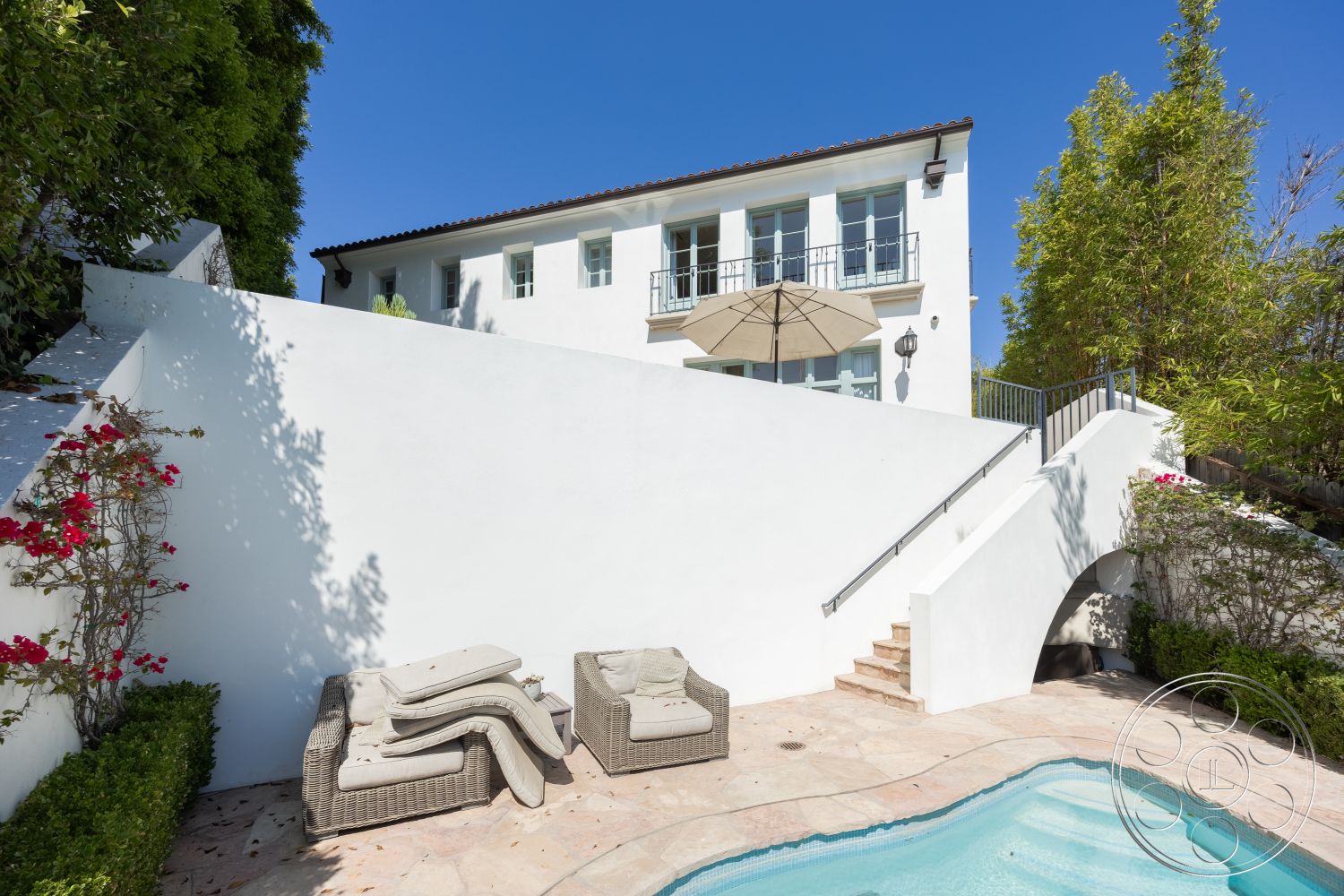 Spanish 168 - terracotta roof tiles, rectangle swimming pool, concrete retaining wall, exterior white stucco walls, stone patio flooring, concrete pool border, integrated outdoor seating area, balcony with city skyline view, outdoor umbrella for shade, wrought iron balcony railing, curved pool steps, landscaped garden with flowering plants, arched doorway leading to pool area, built-in outdoor lighting fixtures, paver walkway leading to pool, fencing with greenery for privacy