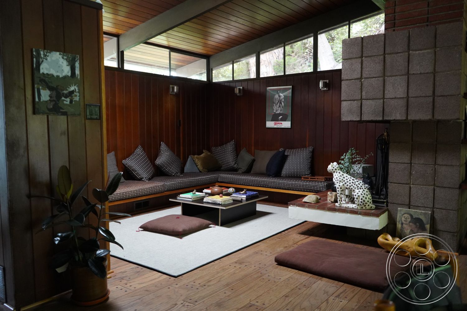 MCM 25 - dark wood paneling interior, exposed wood ceiling beams interior, indoor plant decor interior, wooden flooring interior, cozy reading nook interior, sliding glass windows interior, built-in seating area interior, rectangular coffee table interior, textured area rug interior, polka dot throw pillows interior, multicolored art prints on walls interior, stone fireplace surround interior, natural light-filled living space interior, open layout living space interior, mid-century modern architectural style interior