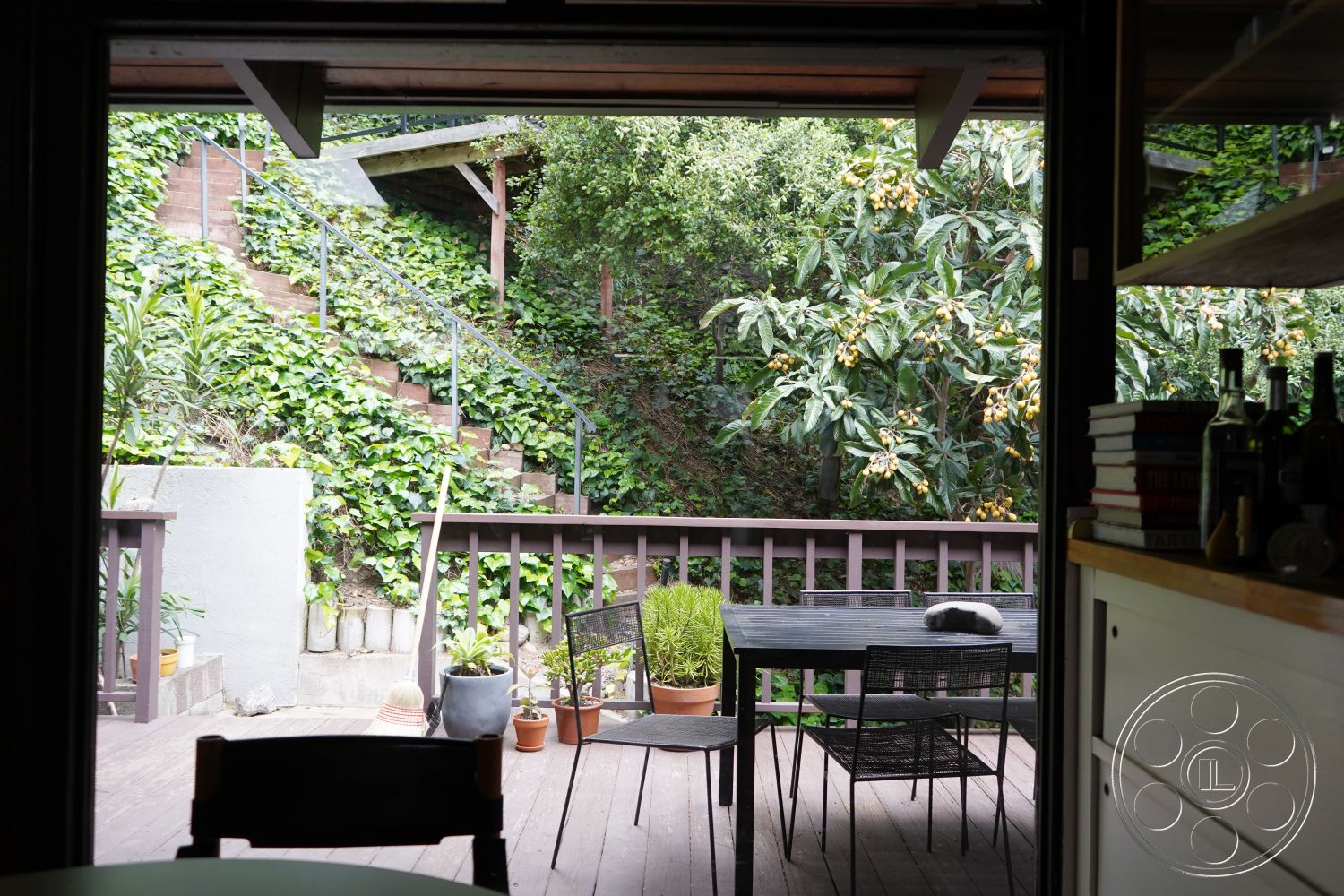 MCM 25 - modern architectural style, natural light from large windows, outdoor dining set, staircase with wooden steps, open-air seating area, concrete retaining wall, wood deck flooring, potted plants on patio, view of landscaped garden, railings with metal spindles, terraced garden with stepping stones, lush greenery surrounding patio area, sloped hillside, interior kitchen view, exterior garden landscape