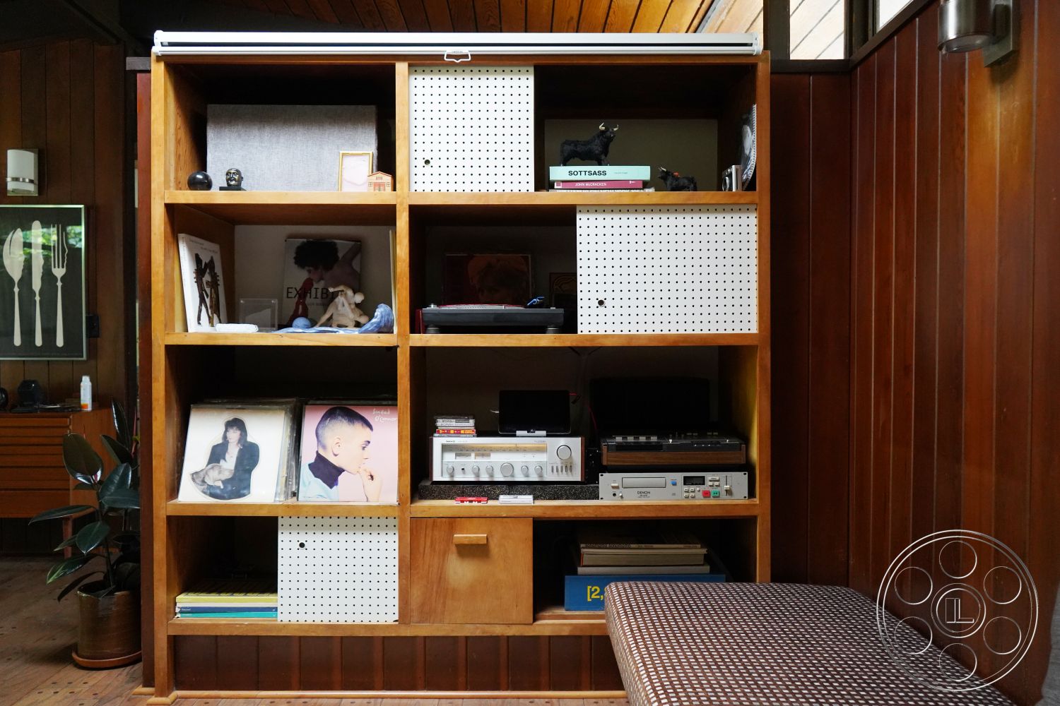 MCM 25 - hardwood floor, natural light from windows, cozy reading nook, retro record player, exposed wood paneling, interior wood shelving unit, mid-century modern bookshelf, open cubby storage design, polka dot fabric drawer, art pieces displayed on shelves, decorative accessories on shelves, vintage audio equipment, photographic prints on display, planted foliage in corner, textured seat cushion