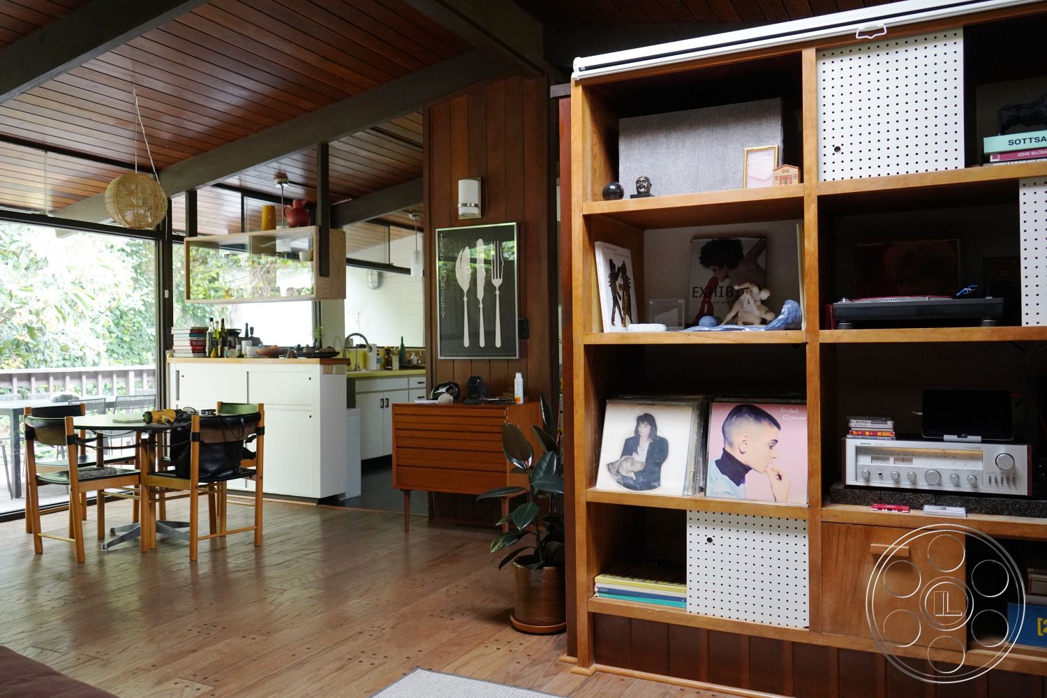 MCM 25 - glass pendant lighting, mid-century modern architectural style, natural light-filled space, white painted walls interior, open floor plan living area, wood ceiling beams interior, vinyl flooring dining area, glass window walls interior, modern wooden furniture, minimalist kitchen island with open shelving, exposed wood cabinets, retro-style record player interior, geometric shelving units interior, green indoor plants interior, city garden view, light wood accents interior