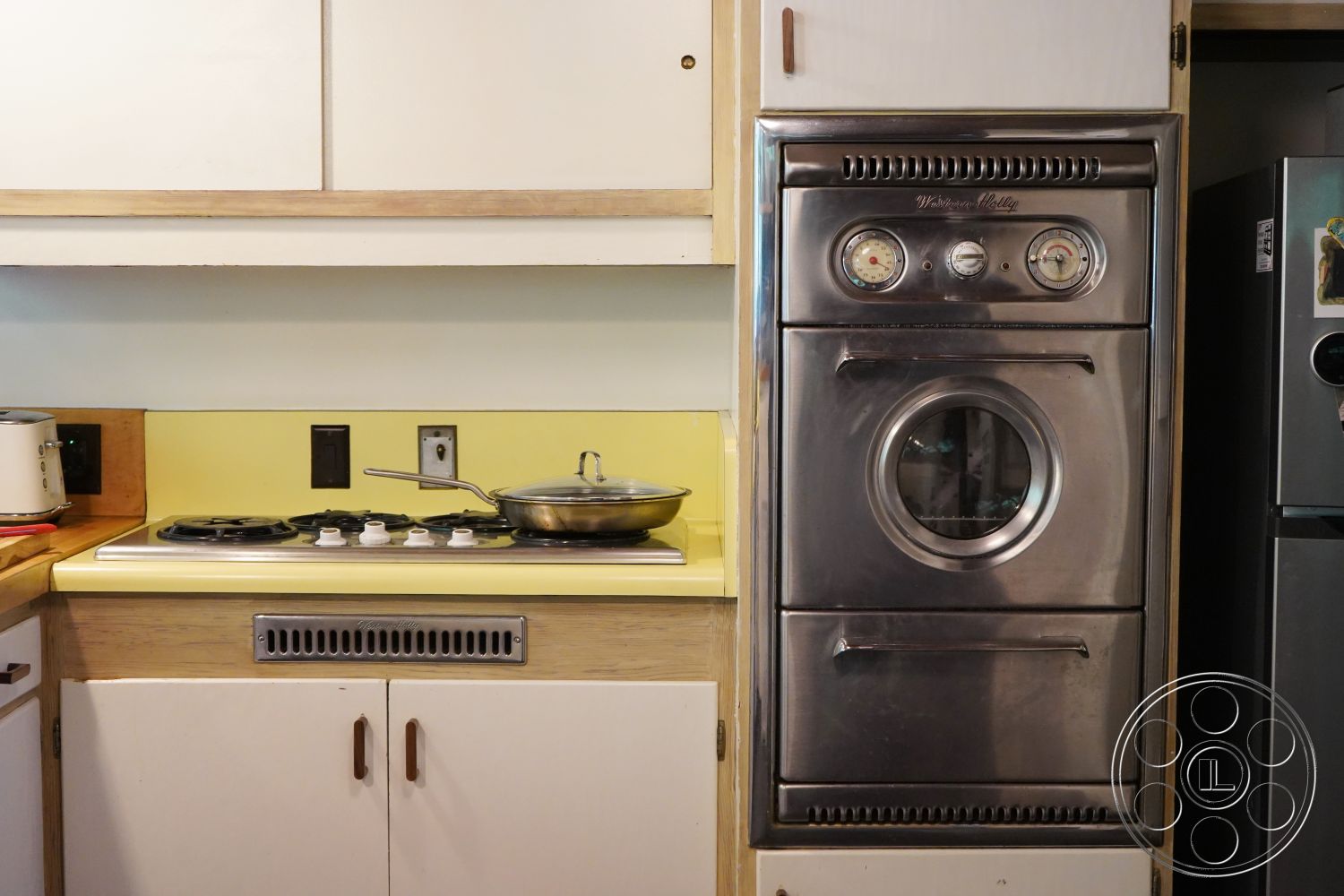 MCM 25 - interior kitchen space, compact kitchen layout, yellow laminate kitchen countertop, stainless steel gas cooktop, chrome vintage oven, white cabinetry with wood accents, black electrical outlets on wall, wooden kitchen cabinet doors, metal kitchen appliance, retro kitchen design