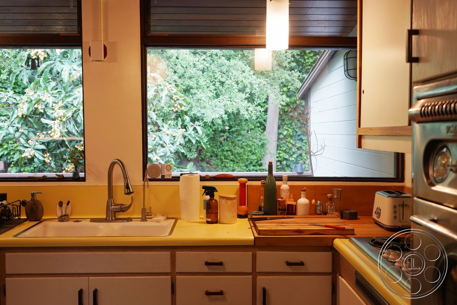 MCM 25 - black faucet with pull-down sprayer, stainless steel appliance finishes, white painted kitchen cabinets, interior kitchen with yellow laminate countertops, dual-pane windows with dark wood trim, wood cutting board on countertop, ceramic dishware on open cabinetry, glass bottle decor on countertops, neutral-colored paper towel holder, modern pendant light fixtures above sink, views of lush greenery outside, hardwood floor under kitchen area, open shelving above kitchen sink, spacious kitchen layout with ample natural light, contemporary kitchen design with functional style