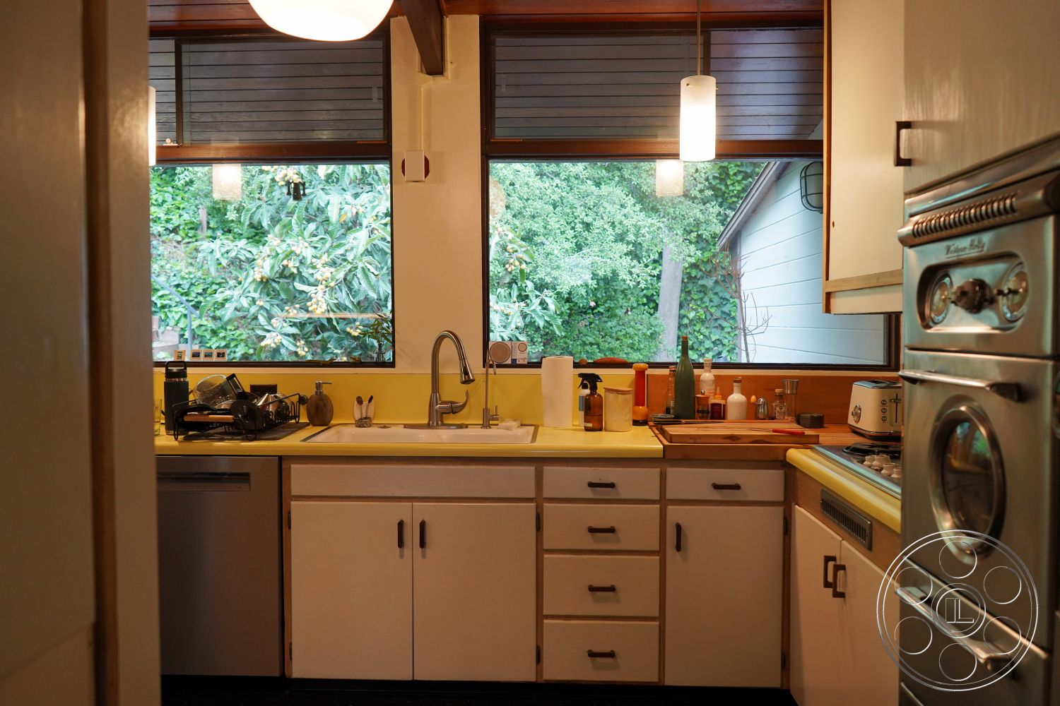 MCM 25 - stainless steel appliances, modern pendant lighting, urban garden view, interior kitchen space, open kitchen layout, minimalist kitchen design, built-in storage cabinets, wooden kitchen flooring, yellow laminate countertops, white cabinetry with simple hardware, double basin sink with faucet, large windows with wooden frames, park-like view through windows, cooking area with gas stove, kitchen utensils and cookware, natural light-filled kitchen