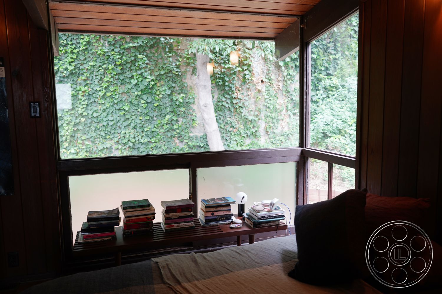 MCM 25 - contemporary design elements, exposed wood ceiling beams, natural light from large window, wood paneling interior, large window with nature view, built-in bookshelf with stacked books, soft bedding with decorative pillows, textured fabric throw blanket, greenery backdrop from outdoor ivy, minimalistic decor with functional furniture, warm wooden finishes, open space bedroom design