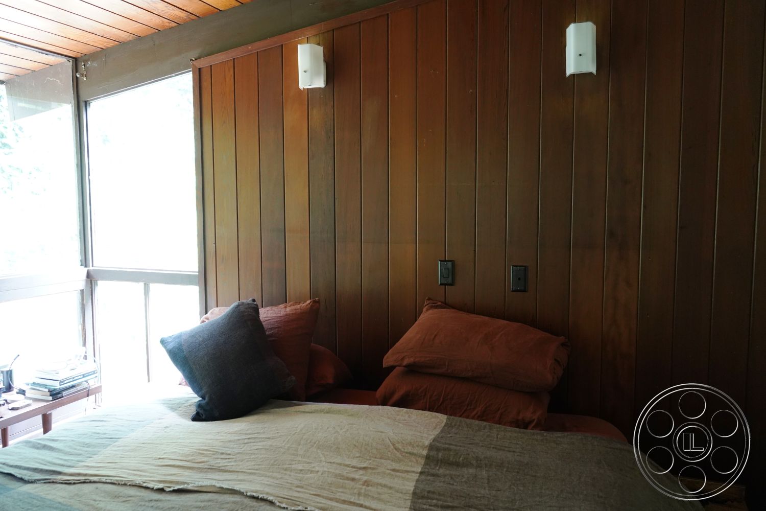 MCM 25 - wooden bedside table, earthy color palette, textured wall treatment, dark wood accent wall, open room layout, large windows with natural light, contemporary design details, interior wooden panel walls, pine wooden ceiling, modern rustic bedroom, minimalist bedding, soft fabric throw pillows, bedside reading lamp, cozy bedroom seating area, natural setting view