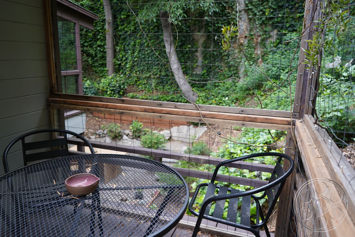 MCM 25 - natural landscaping, contemporary outdoor dining area, wooden deck railing, forest setting, shaded outdoor space, powder-coated steel patio table, metal mesh patio chairs, exposed wood structure, green leafy garden view, mesh garden enclosure, wooden vertical railing, enclosed outdoor patio, wooden board fence, interior patio area