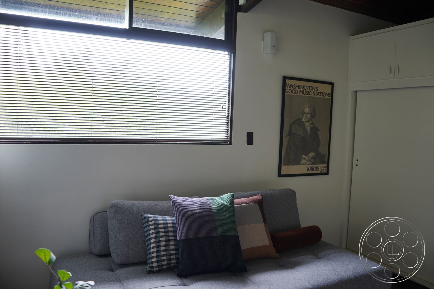 MCM 25 - natural light, open floor plan, minimalistic decor, colorful throw pillows, slatted window blinds, gray fabric sofa, neutral painted walls, built-in storage cabinets, wood beam ceiling, indoor potted plant, large sliding glass windows, exposed wood wall panel, framed vintage wall art, contemporary living area