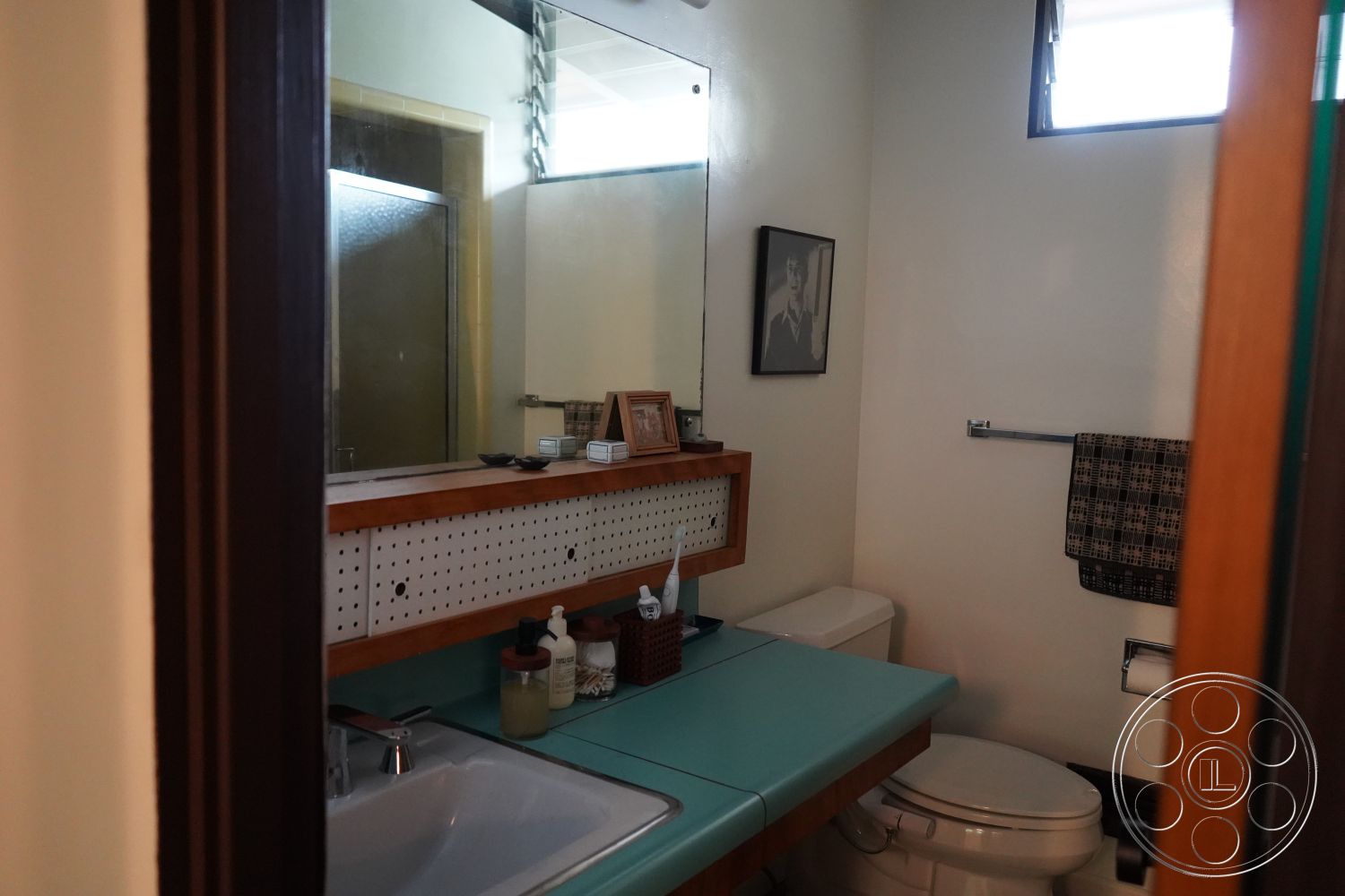 MCM 25 - polished concrete flooring, natural light from window, interior bathroom, walnut vanity with perforated paneling, turquoise laminate countertop, modern sink with chrome faucet, white ceramic toilet, black and white framed artwork, framed mirror with minimalist design, textured shower wall, half-tiled wall, window with frosted glass, ceramic soap and lotion dispenser, black and white patterned towel, functional and stylish storage options