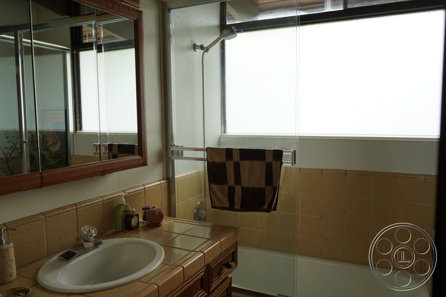 MCM 25 - neutral color palette, white porcelain sink, under-mounted sink, glass shower doors, modern bathroom fixtures, recessed lighting fixtures, natural light from large window, minimalist bathroom design, natural wood bathroom cabinetry, interior bathroom, ceramic tile bathroom flooring, mirrored medicine cabinet, wood-framed bathroom mirror, beige tiled bathroom walls, frosted glass window, shower area with glass partition, contemporary bath rug, towel bar with decorative towels