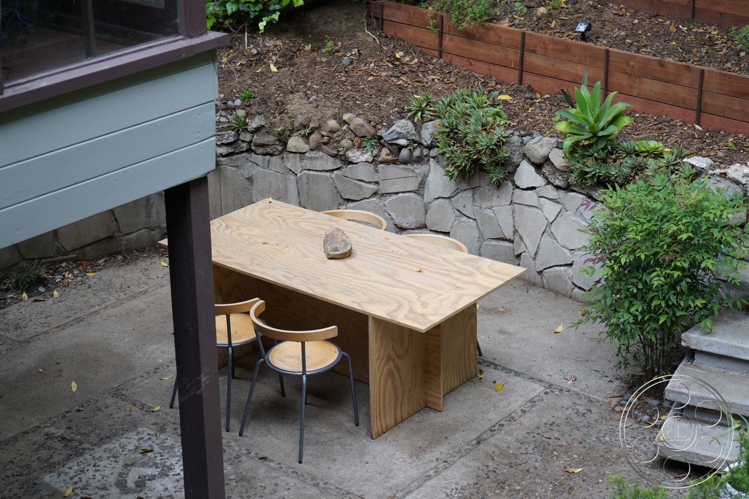 MCM 25 - surrounding greenery, outdoor dining area, stepping stone pathway, natural stone retaining wall, metal dining chairs, plywood dining table, patio area exterior, rough stone wall, wooden garden bed borders, landscaped garden plants, textured concrete slab floor, earthy tones in design