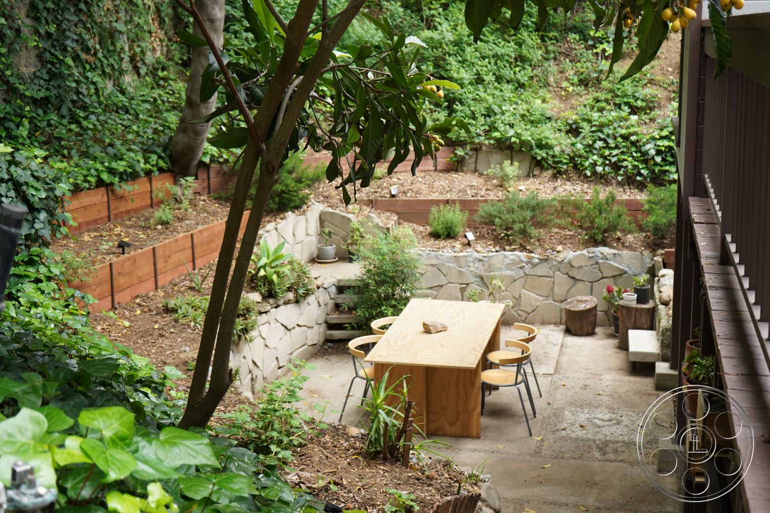MCM 25 - outdoor dining space, natural stone wall, urban backyard, metal dining chairs, landscaped garden area, exterior garden patio, lush greenery setting, surrounding trees and shrubs, stone patio flooring, wooden garden table, retaining wall with wood paneling, textured earth surface, sunken garden design, mixed landscape textures, low-maintenance garden design, container gardening elements