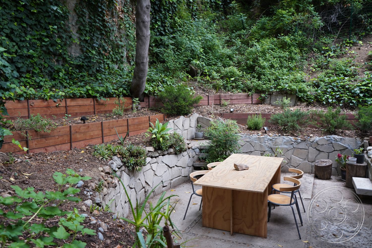 MCM 25 - exterior, intimate dining space, environmentally integrated design, natural stone retaining wall, earth-tone color palette, natural wood texture, metal dining chairs, lush greenery setting, wooden raised garden beds, concrete patio floor, urban backyard retreat, minimalist outdoor dining table, surrounding hillside vegetation, decorative stone features, functional garden landscaping, relaxed outdoor aesthetic