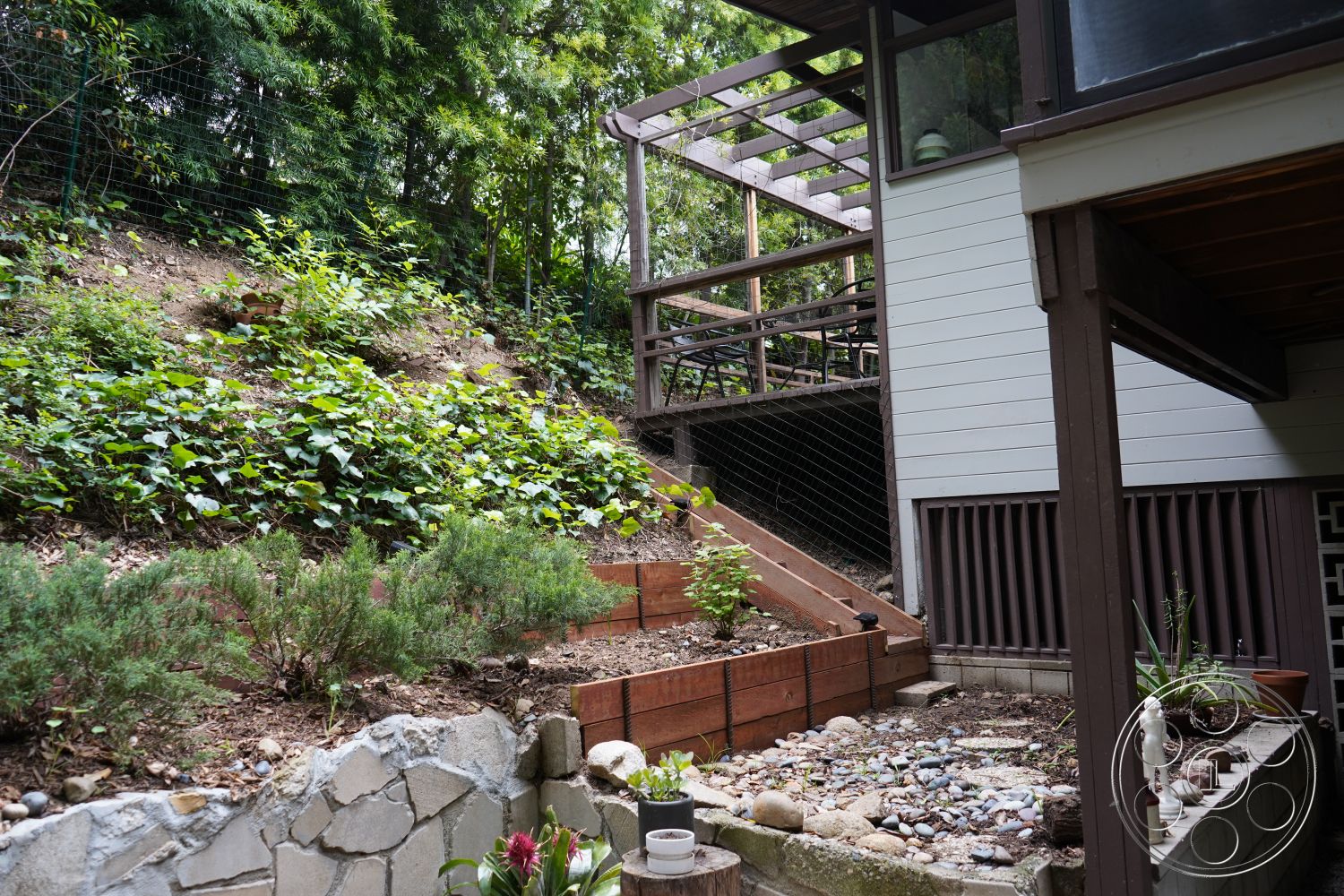 MCM 25 - wooden planter boxes, garden patio, natural stone pathway, outdoor living area, concrete retaining wall, natural landscape setting, stone garden wall, lush greenery landscaping, interior outdoor space, wooden deck stairs, exposed wood decking, gravel landscaping ground cover, sloped hillside garden, wood trellis structure, mixed vegetation garden, rear yard, urban hillside property