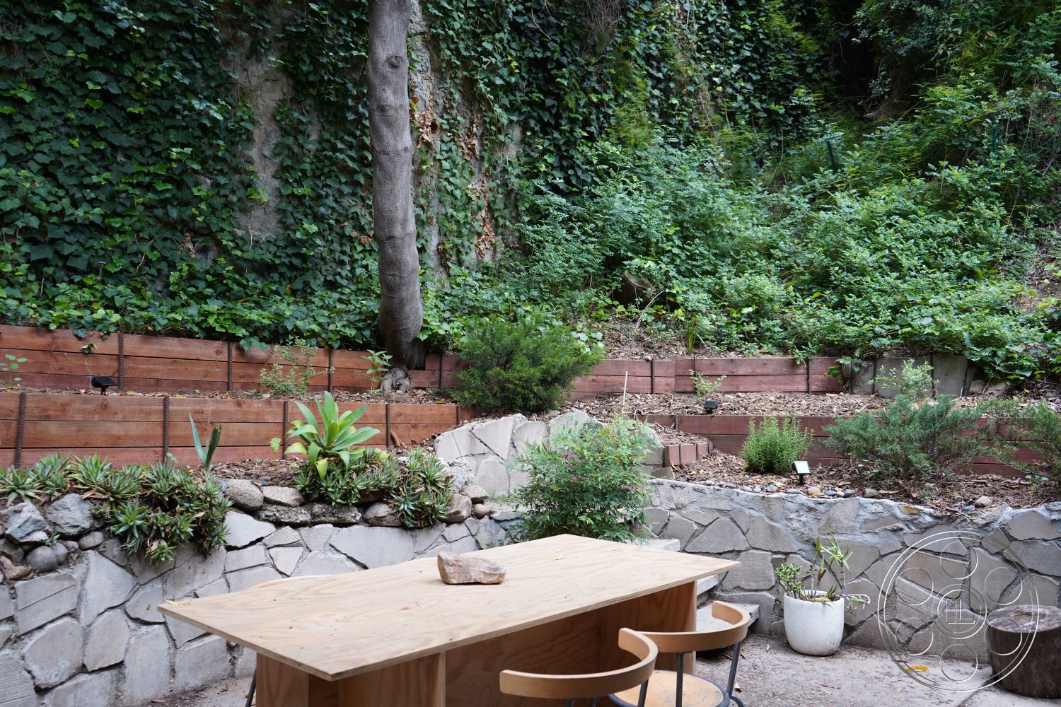 MCM 25 - exterior shaded seating area, exterior horizontal wood slats, exterior wooden dining table, exterior lush greenery backdrop, exterior potted plants, exterior stone wall garden bed, exterior natural stone landscaping, exterior wooden retaining walls, exterior natural earth tones, exterior rustic garden design, exterior tiered garden layout, exterior natural light dining area, exterior organic materials, exterior informal outdoor setting