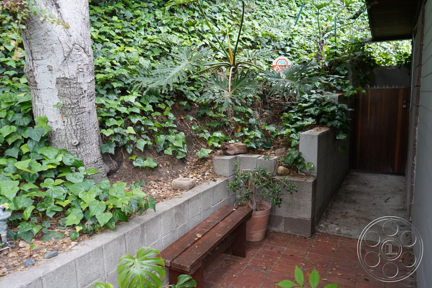 MCM 25 - natural stone accents, wooden bench seating, green foliage backdrop, concrete block garden wall, exterior garden area, brick patio flooring, native plant landscaping, low-maintenance garden design, irregular stone pathway, rustic wooden privacy gate, textured earth tones, vertical garden wall, sloped garden bed, suburban walkway, lush greenery environment