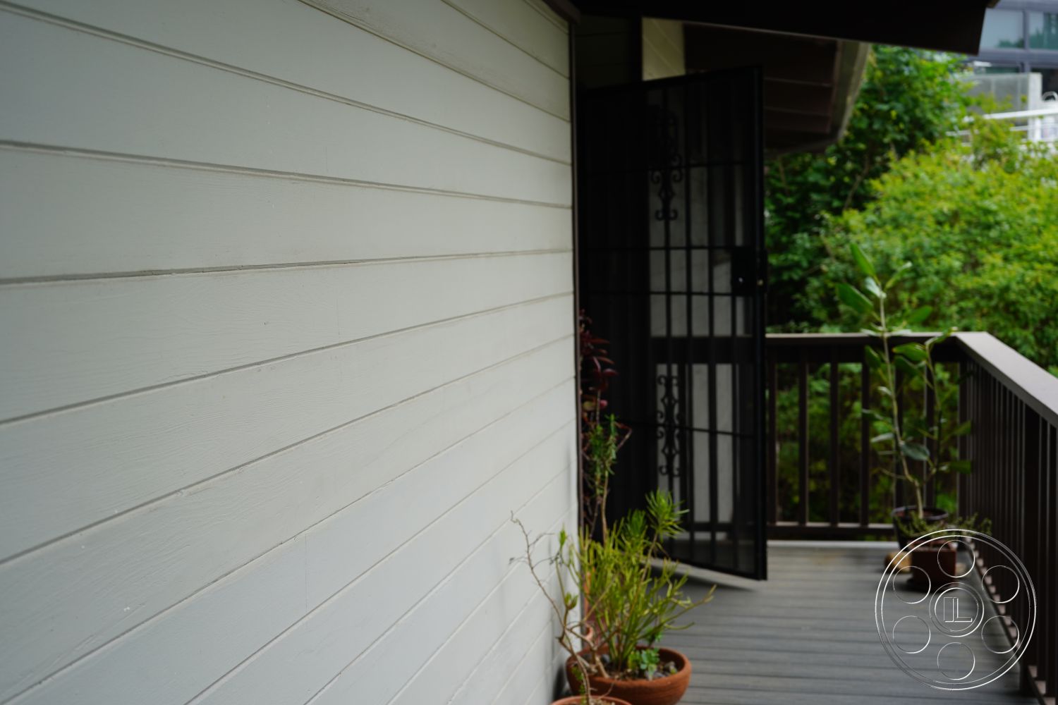MCM 25 - landscaped garden setting, minimalist architectural design, flat roof overhang, natural light from outdoor view, exterior wood siding wall, painted wood deck flooring, metal security screen door, outdoor planters with greenery, curved rail balcony, contemporary outdoor entryway