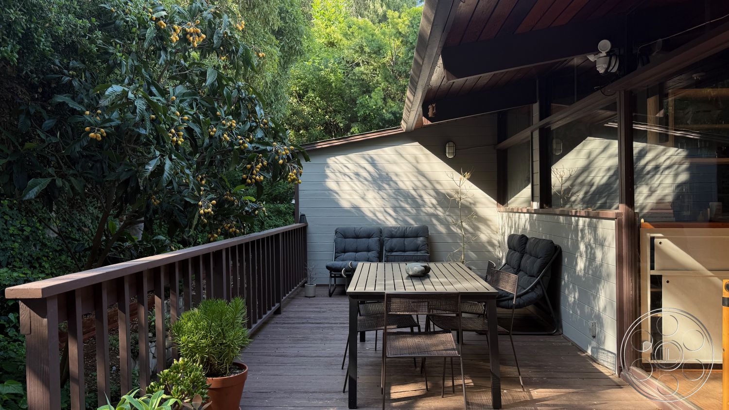 MCM 25 - modern outdoor furniture, floor-to-ceiling windows, outdoor dining area, shaded seating area, relaxed outdoor ambiance, natural wood finish, lush green landscaping, gabled roof design, urban garden setting, integrated indoor-outdoor living, wooden deck patio, textured wood railing, exterior wall with grey wood siding, tropical plants surrounding