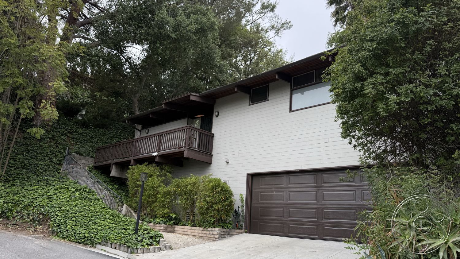 MCM 25 - exterior, concrete driveway, surrounded by greenery, natural stone pathway, two-story home, ranch-style home, landscaped garden with shrubs, wood siding exterior, sloped hillside setting, large front windows, brown wooden balcony railing, wooden front steps, integrated garage door, minimalistic architectural style