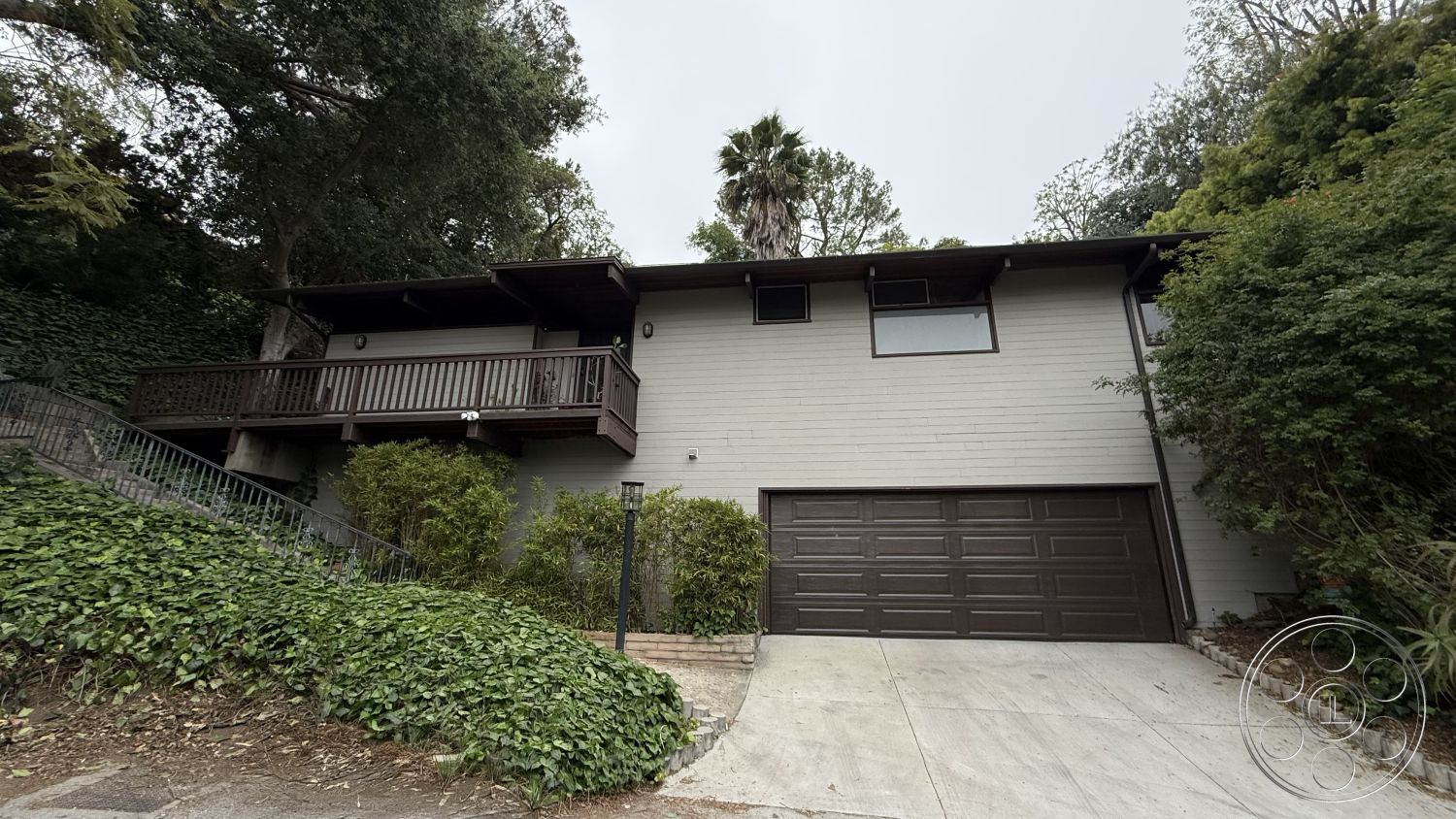 MCM 25 - surrounding greenery, modern architectural style, concrete driveway, sloped driveway, hillside location, paved stone pathway, two-story home, gabled roof design, exterior recessed lighting, horizontal wood siding exterior, wooden balcony railing, landscape garden setting, single car garage, natural wooden supports, mature trees in environment