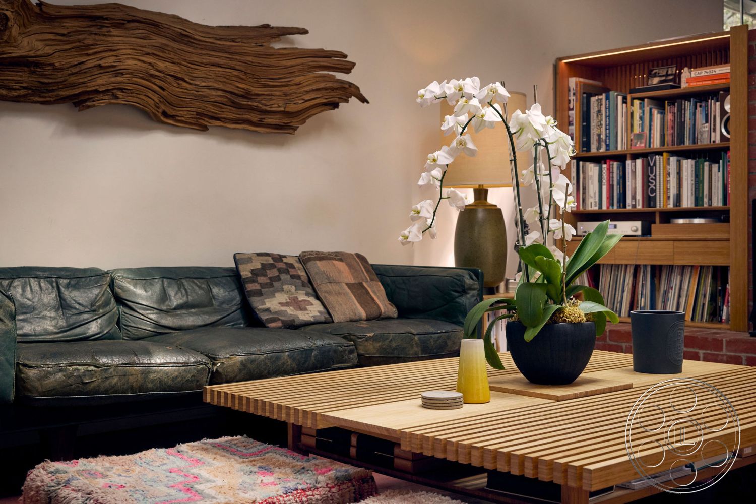 MCM 26 - open living space interior, warm ambient lighting interior, leather sofa seating interior, textured wood wall art interior, unpolished hardwood coffee table interior, brick wall accent interior, tile flooring interior, decorative orchids plant interior, ceramic pot indoor plants interior, built-in wooden shelving unit interior, polyester rug interior, glass lampshade floor lamp interior, geometric patterned throw pillows interior, mid-century modern design interior, natural materials aesthetic interior