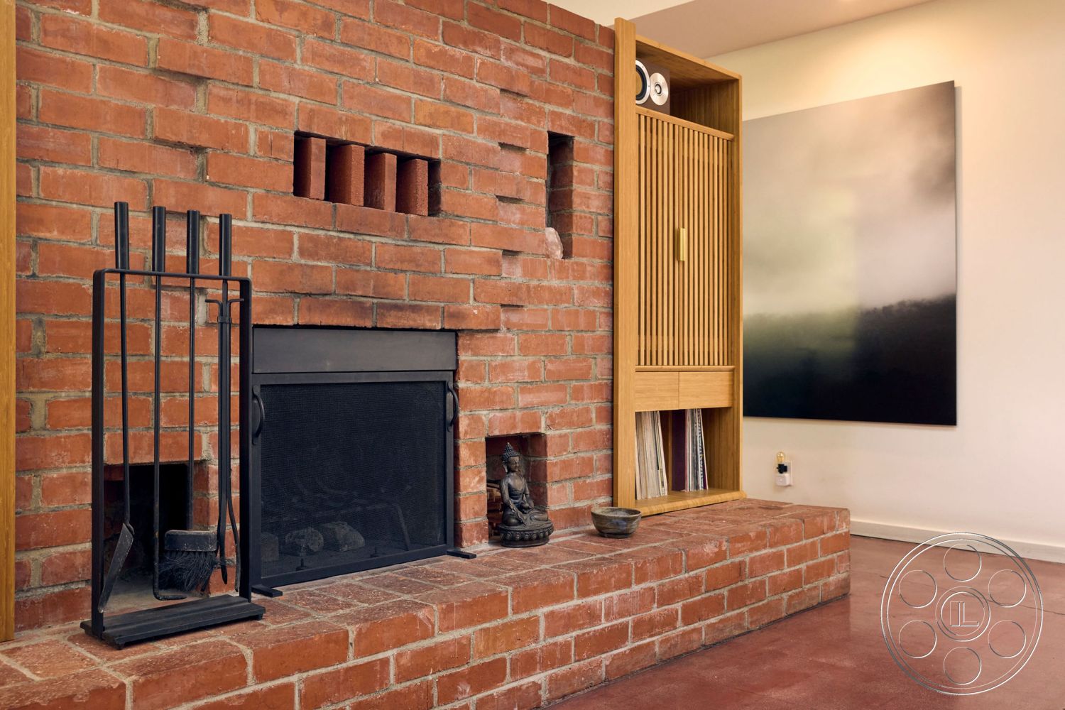 MCM 26 - polished concrete floor, earthy color palette, exposed brick fireplace wall, rustic modern aesthetic, open interior layout, functional living space, brick hearth with built-in wood storage, black metal fire screen, light wood cabinetry with slatted doors, minimalistic modern art wall decor, warm-toned wood accents, contemporary living room setting, cozy fire feature, detailed craftsmanship in cabinetry