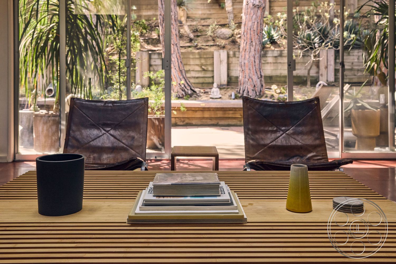 MCM 26 - interior minimalist decor, interior hardwood flooring, exterior landscaped garden view, interior open layout, interior low-profile coffee table, interior potted greenery, interior glass sliding doors, interior warm color palette, interior leather lounge chairs, exterior tall pine trees, exterior stone garden walls, interior wooden slat dining table, interior black ceramic planter, interior natural light from glass doors, interior stacked books and decorative items on table