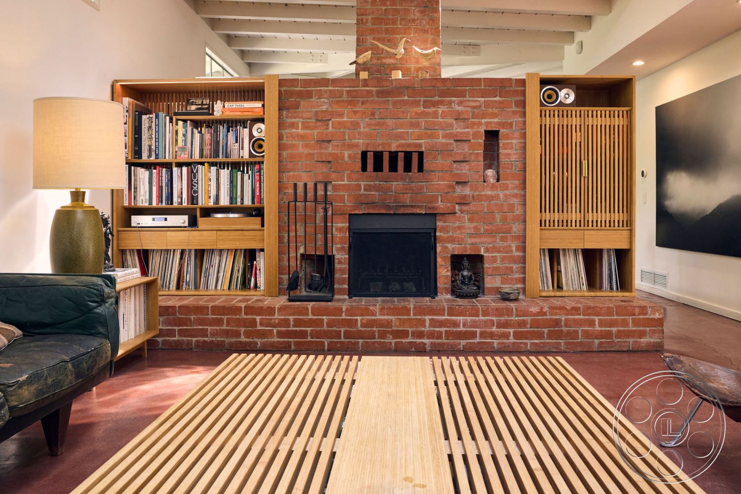 MCM 26 - light wood cabinetry, natural light from large windows, exposed wood ceiling beams, mid-century modern architectural style, cozy living room setting, exposed brick accent wall, modern recessed lighting, textured plaster interior walls, minimalist floor lamp, wooden built-in bookshelves, contemporary wooden coffee table, tile flooring with reddish tone, stacked brick fireplace surround, open living area layout, stylish chair with metal legs