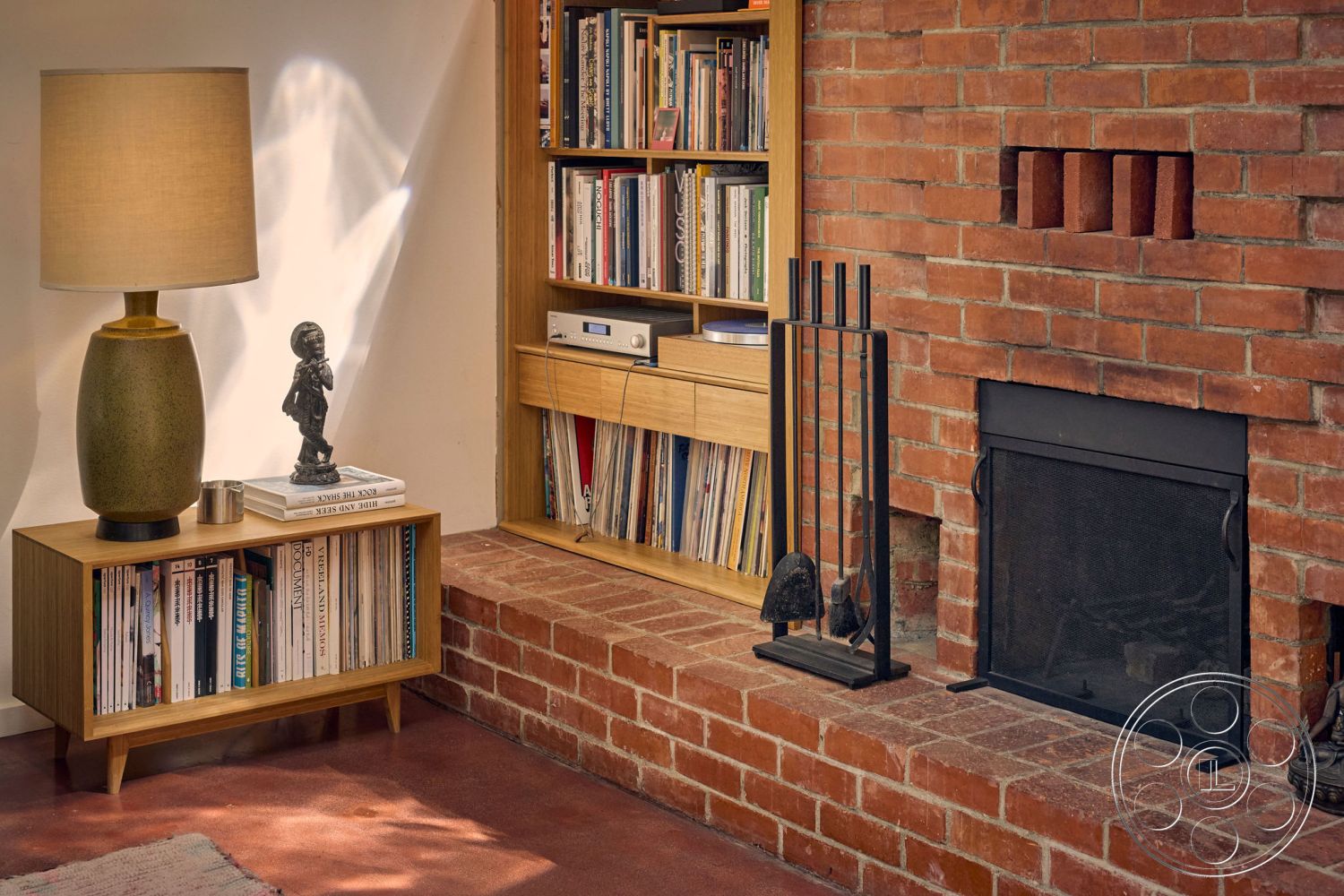 MCM 26 - warm ambient lighting, wooden media console, exposed brick fireplace wall, cozy living room setting, textured plaster interior walls, brick hearth, wooden bookshelf with built-in storage, contemporary lamp with fabric shade, dark metal fireplace screen, hardwood floor in living area, mid-century modern furniture style, decorative statue on console