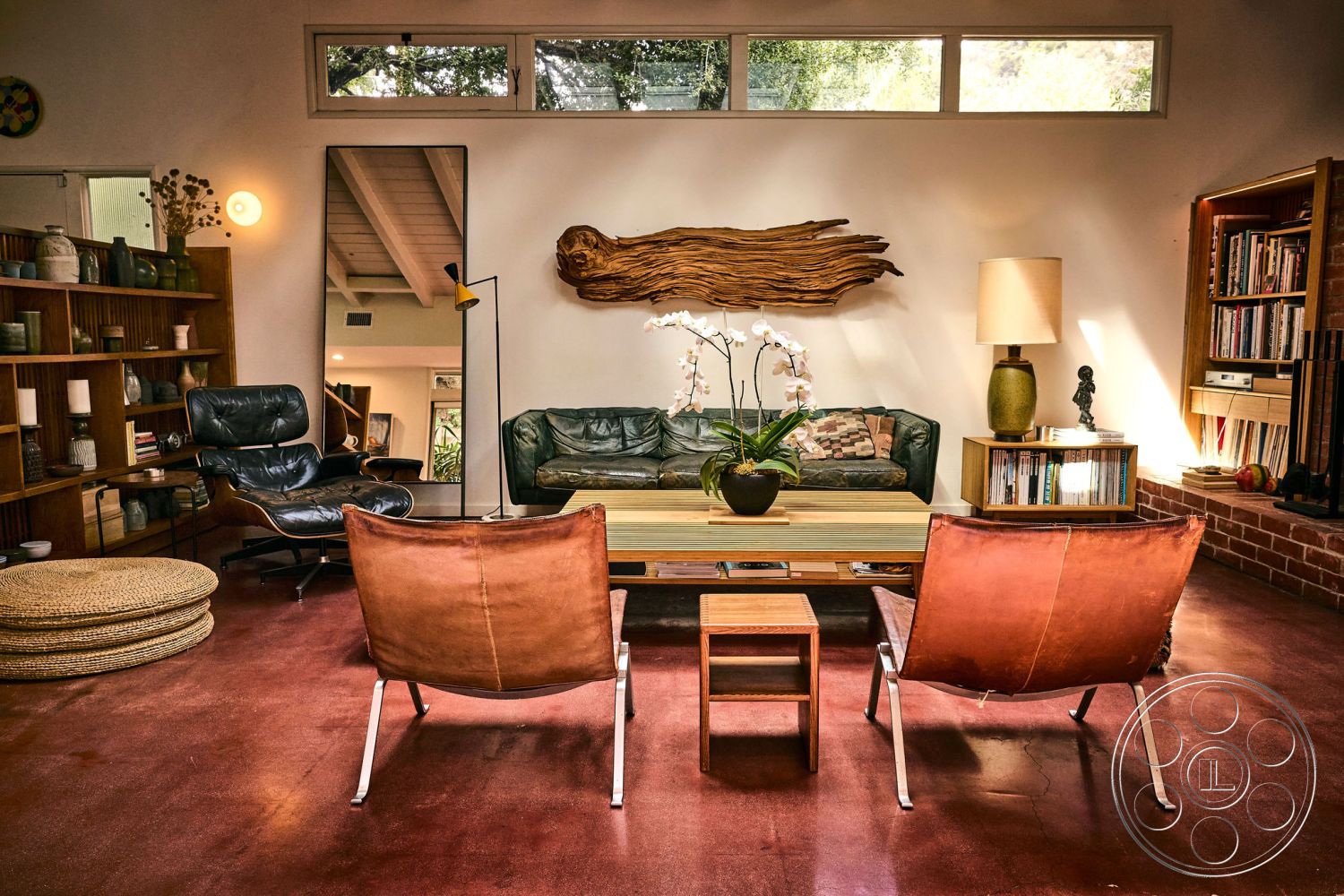 MCM 26 - wooden coffee table, polished concrete flooring, brick accent wall, exposed wood ceiling beams, floor-to-ceiling windows for natural light, leather lounge chairs, green leather sofa, interior mid-century modern living room, plant decor with orchids, open bookshelf with decorative items