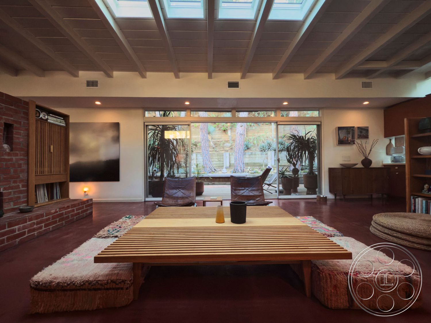 MCM 26 - wood beam ceiling interior, open floor plan interior, indoor plant decor interior, sliding glass doors interior, textured area rug interior, mid-century modern design interior, exposed brick fireplace surround interior, red concrete floors interior, natural light with skylights interior, wood slatted coffee table interior, wooden shelving unit interior, dark wood accent furniture interior, minimalist design style interior, garden view through glass doors interior