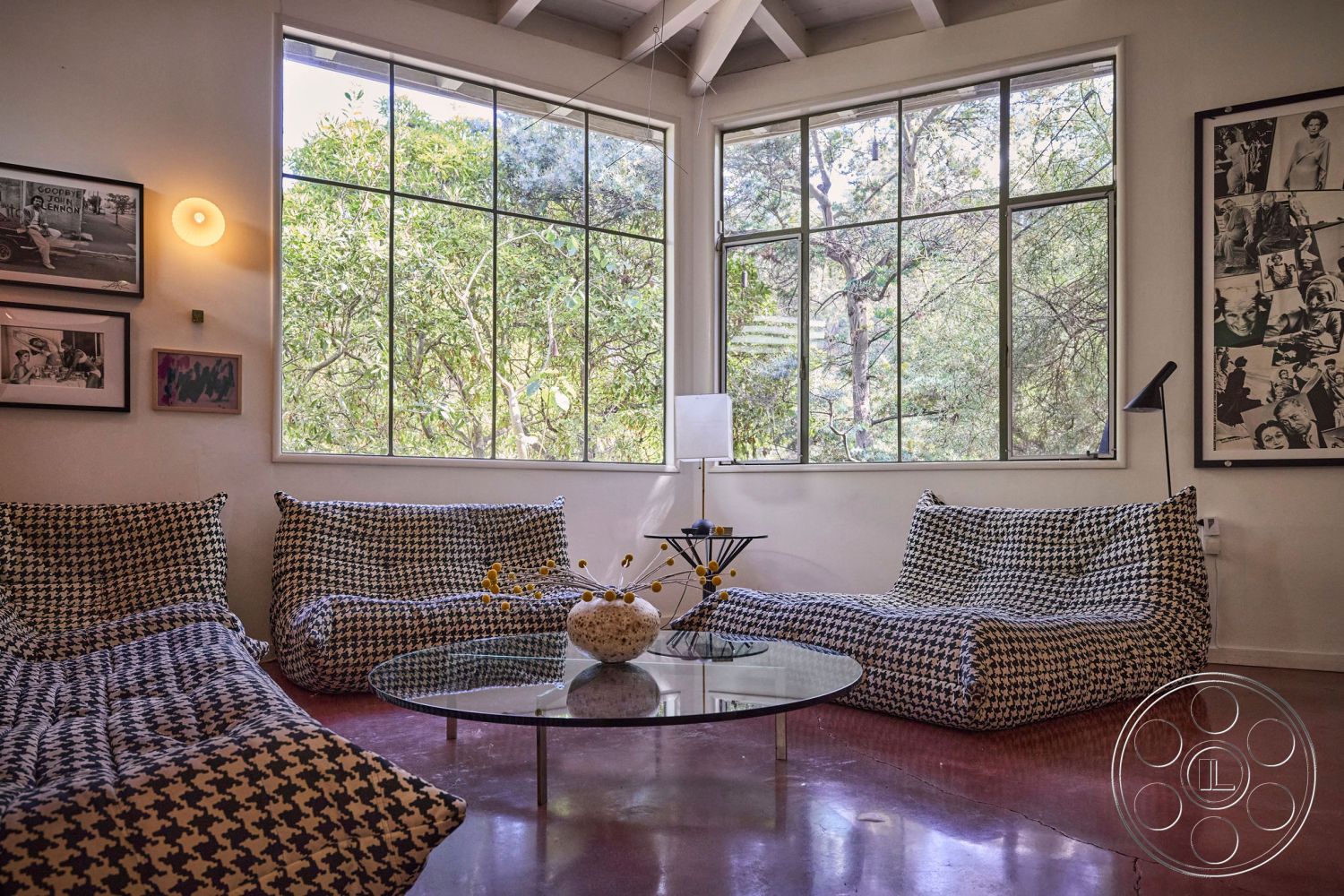 MCM 26 - polished concrete floor, large floor-to-ceiling windows, round glass coffee table, minimalist décor, wall-mounted light fixtures, contemporary lounge area, natural light interior, open layout living space, exposed wooden beams ceiling, black-and-white houndstooth upholstery, urban forest view setting, art deco framed photographs