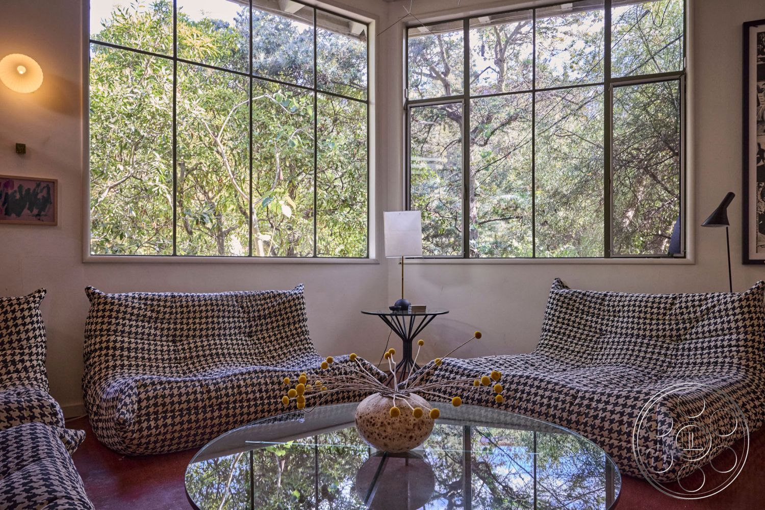 MCM 26 - minimalist decor, modern light fixtures, textured plaster walls, round glass coffee table, interior living room with large windows, contemporary furniture with houndstooth pattern, wooden floor with warm reddish tones, indoor plants visible through windows, natural woodland view, open and airy room layout