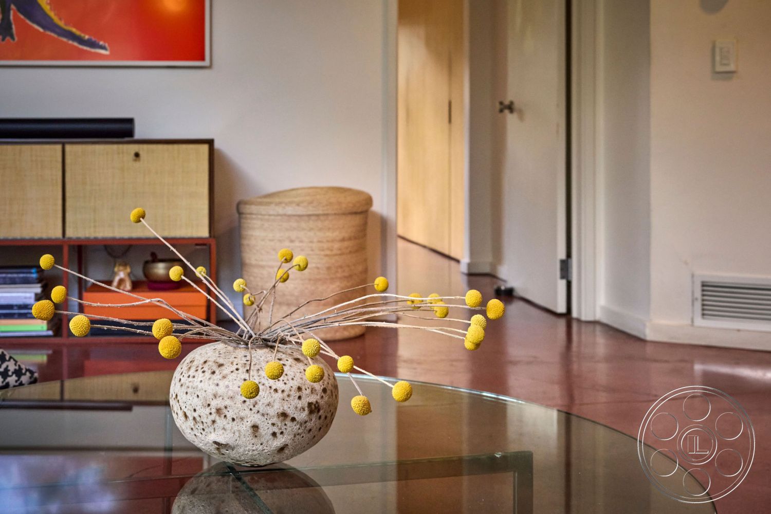 MCM 26 - glass coffee table, interior space, neutral wall color, textured wall treatment, woven storage basket, mid-century modern style, exposed wood ceiling beams, minimalist decor elements, concrete polished floor, simple door design, open floor layout, artwork with vibrant colors, natural light source, decorative vase with yellow accents, modern sideboard with natural finish