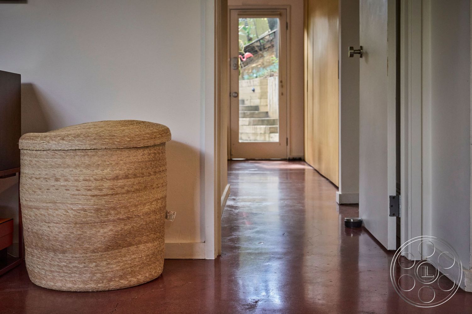 MCM 26 - interior hallway with polished concrete floor, exposed wooden door frame, neutral wall treatment with light paint, woven storage ottoman in a contemporary style, linear hallway layout with indirect lighting, modern aesthetic with minimal furnishings, interior space leading to outdoor stairs, natural light from glass door, smooth transition from indoor to outdoor areas