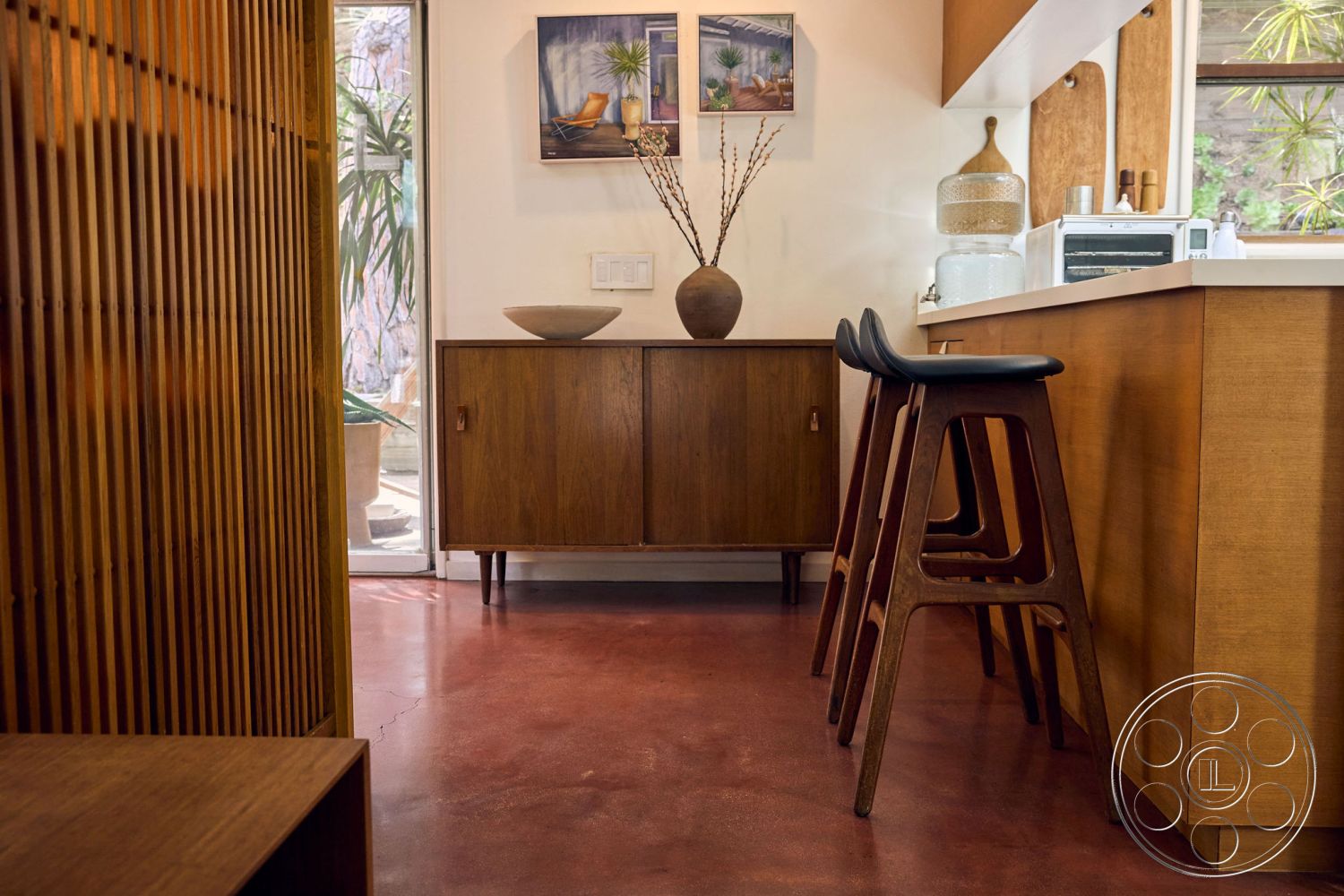 MCM 26 - indoor plants, natural wood accents, polished concrete flooring, minimalist design elements, nature-inspired decor, wall-mounted artwork, open kitchen layout, textured wood wall treatment, mid-century modern cabinetry, stool height seating, bar-height countertop, bright airy space, functional kitchen area, smooth surface countertops