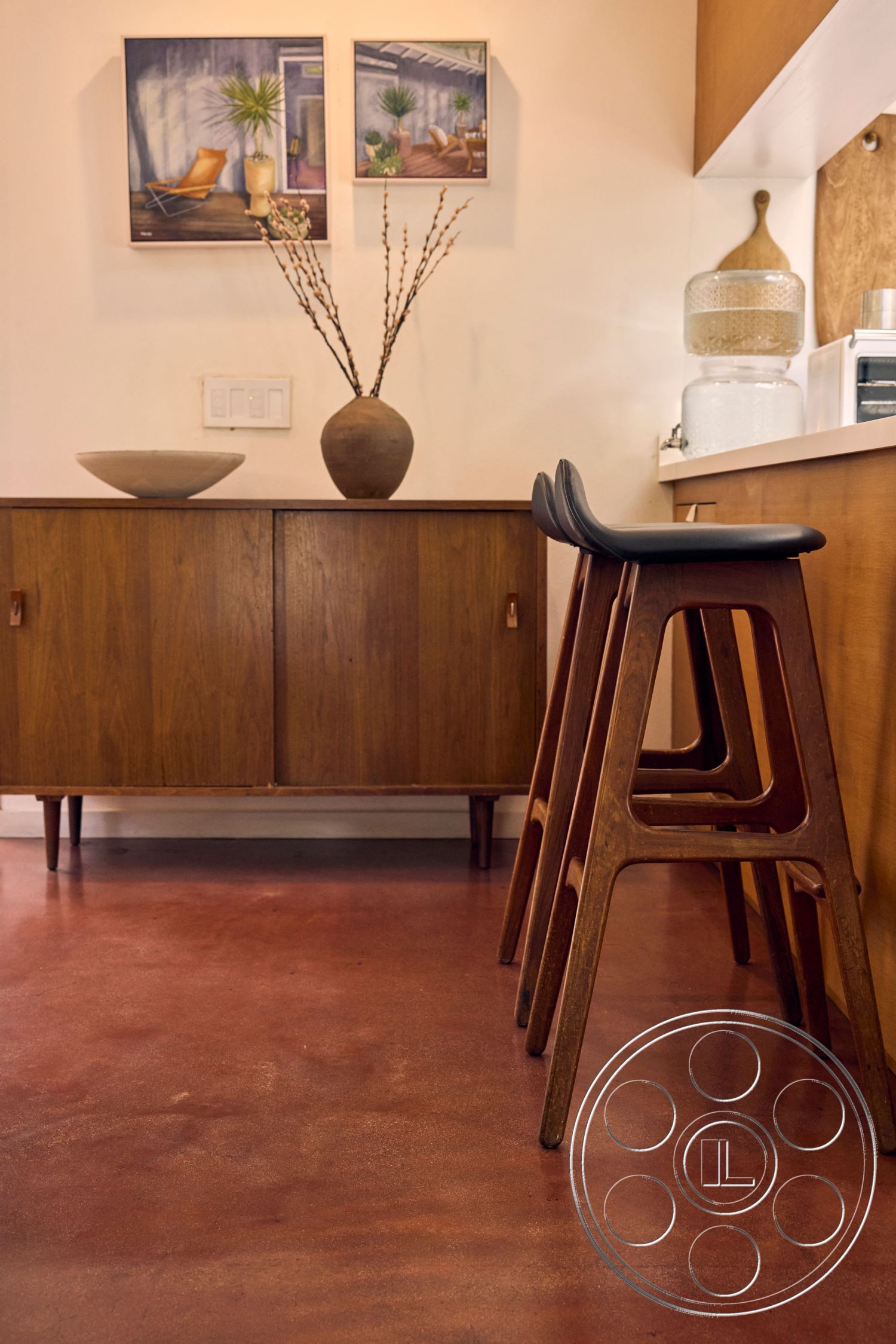 MCM 26 - textured wall art, wooden bar stools, earth-toned color palette, minimalist interior design, interior polished concrete floor, mid-century modern cabinetry, natural wood cabinet, accent decor with ceramic vase, light streaming through window, simple contemporary styling