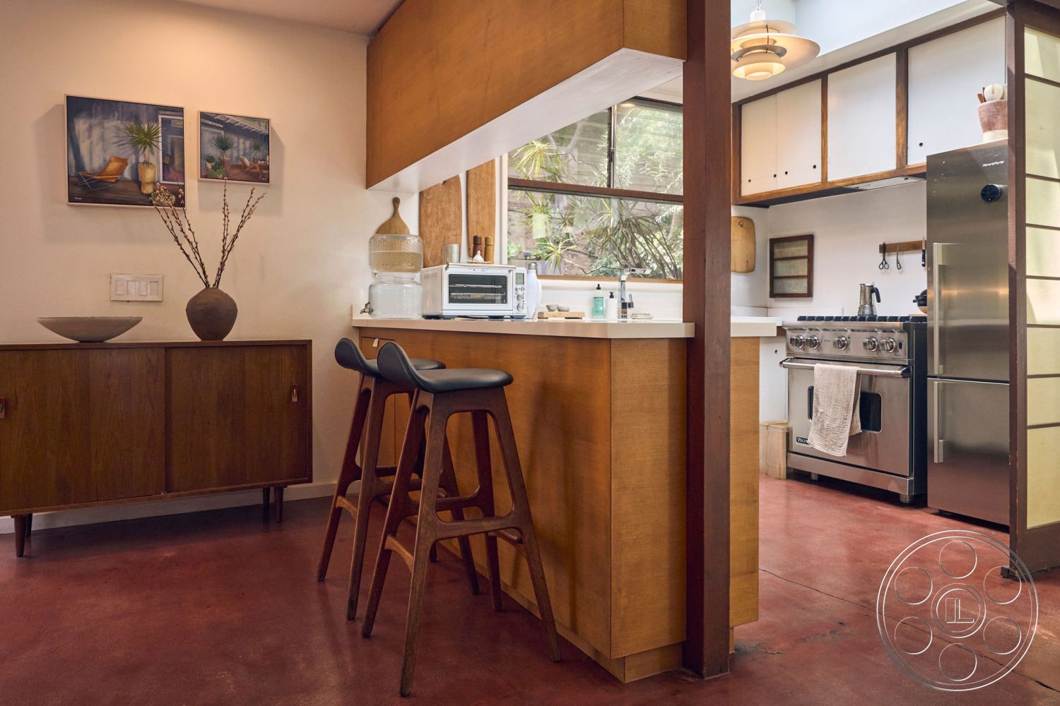 MCM 26 - dark wood accent wall, stainless steel kitchen appliances, mid-century modern architectural style, modern pendant lighting fixtures, decorative plant accents, bright and airy interior, exposed concrete interior floor, wooden cabinetry with minimalist design, white quartz countertop with integrated sink, open kitchen layout with bar seating, two-tone wood and white color scheme, floor-to-ceiling windows allowing for natural light, retro-style bar stools, textured wall treatments with a natural finish, minimalist kitchen backsplash, rustic decor items like vases and bowls, kitchen workspace with minimalist aesthetics, indoor plant setting, stylish kitchen island with seating