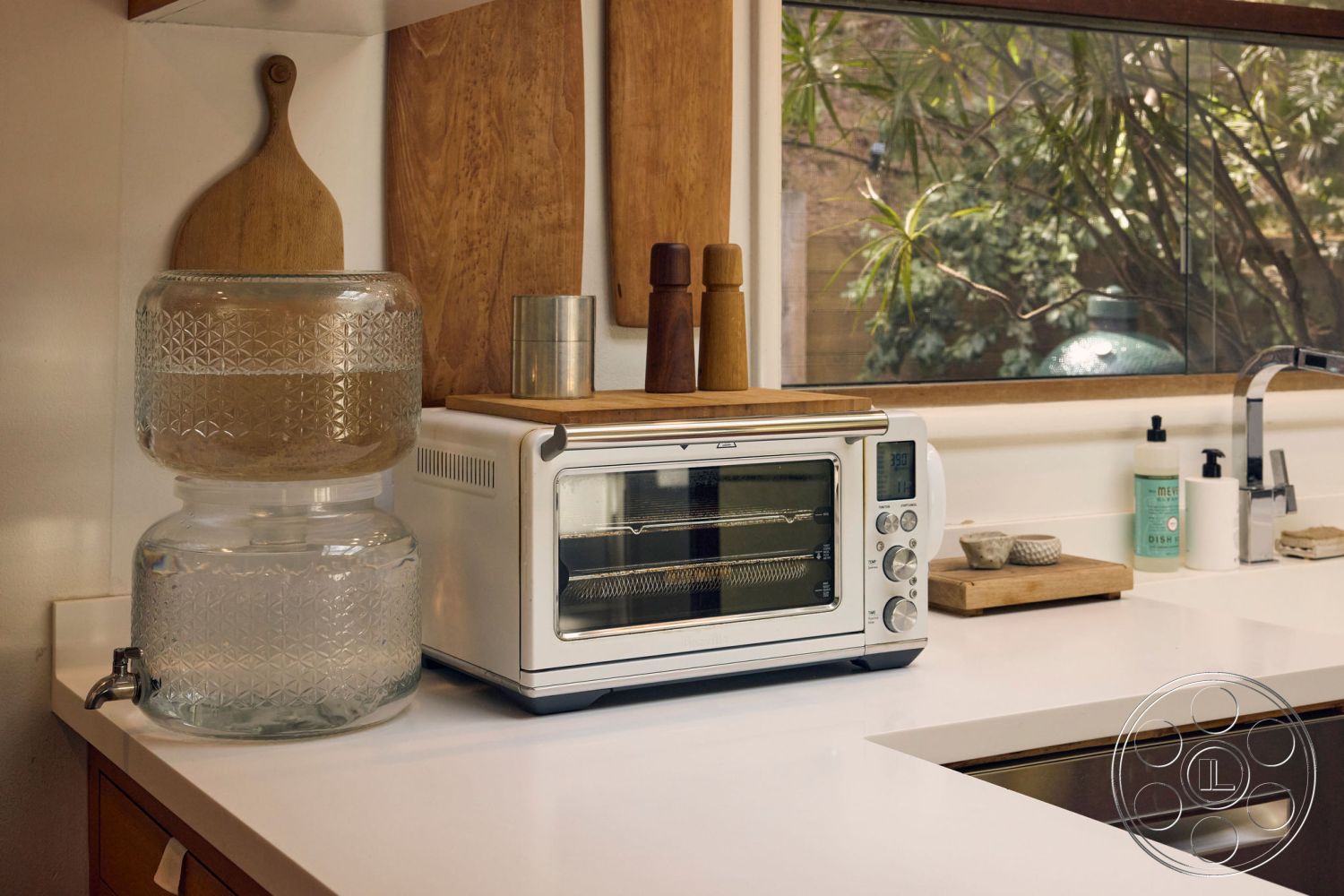 MCM 26 - modern minimalist decor, natural light-filled space, large window with garden view, wood cabinetry with natural finish, wood cutting board on countertop, interior kitchen with white quartz countertop, glass beverage dispenser with pattern detailing, stainless steel appliance with digital display, medium brown wood wall treatment, glass backsplash behind countertop, herb container on kitchen counter